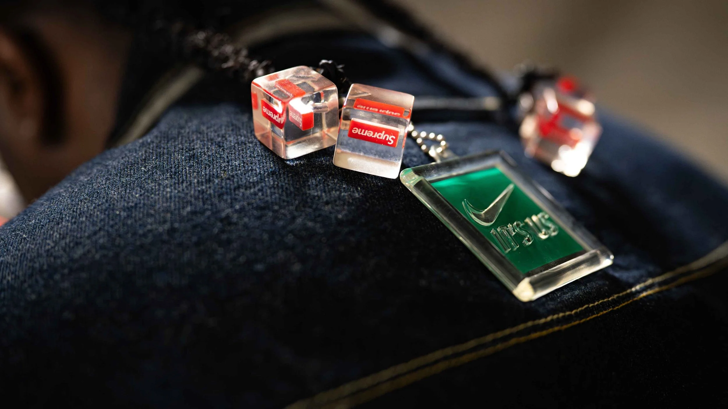 Close-up of denim fabric with a 'YES' badge, two transparent dice with 'Supreme' logos, and a keychain in the shape of a mushroom on the pocket.
