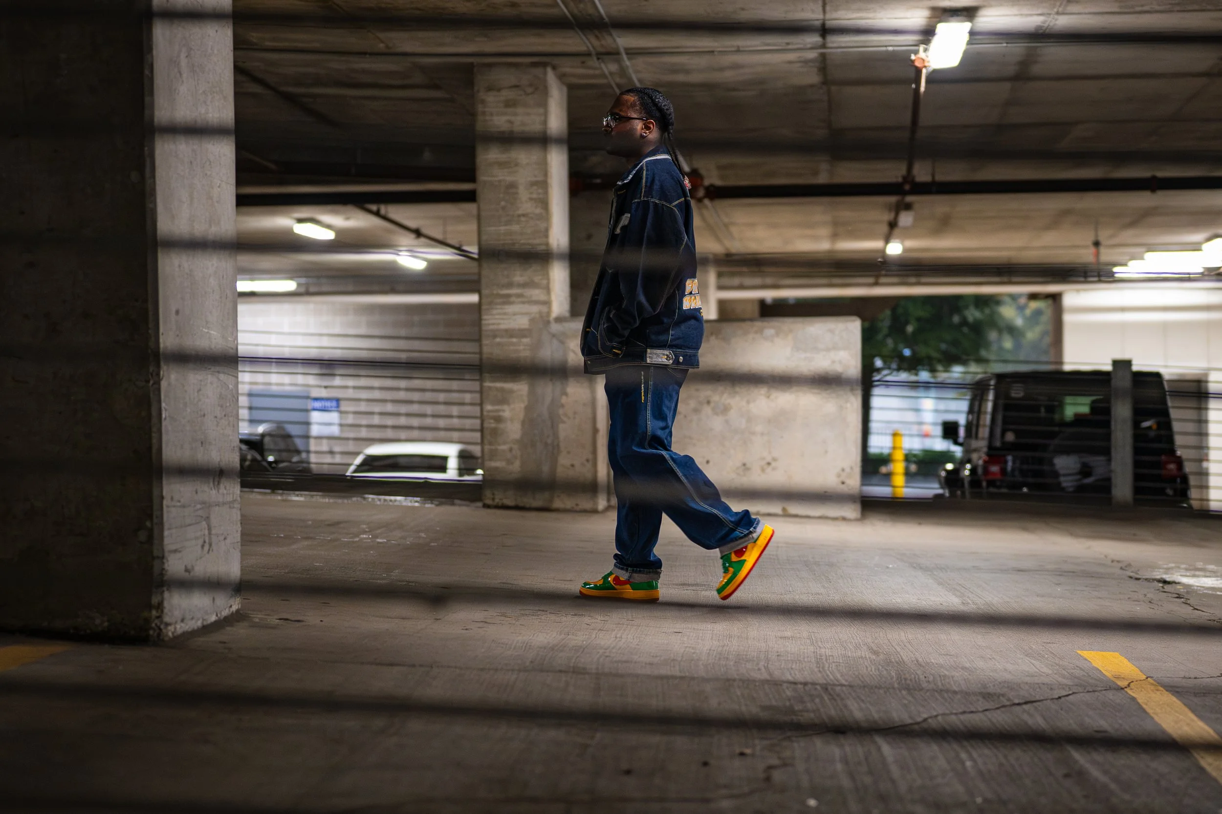 Person walking through a concrete parking garage, wearing a denim jacket, jeans, and bright sneakers.
