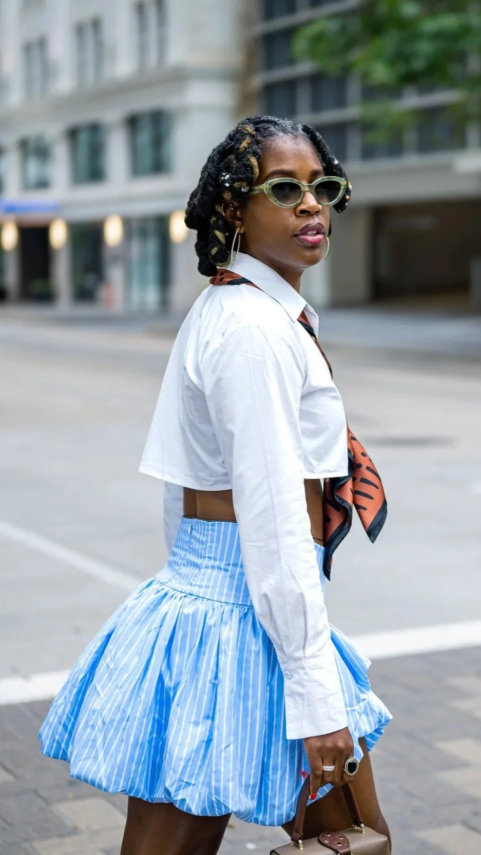 A woman with styled black dreadlocks, wearing large sunglasses, a white cropped shirt, a blue striped ruffled skirt, and a patterned scarf around her neck, standing on an urban street.