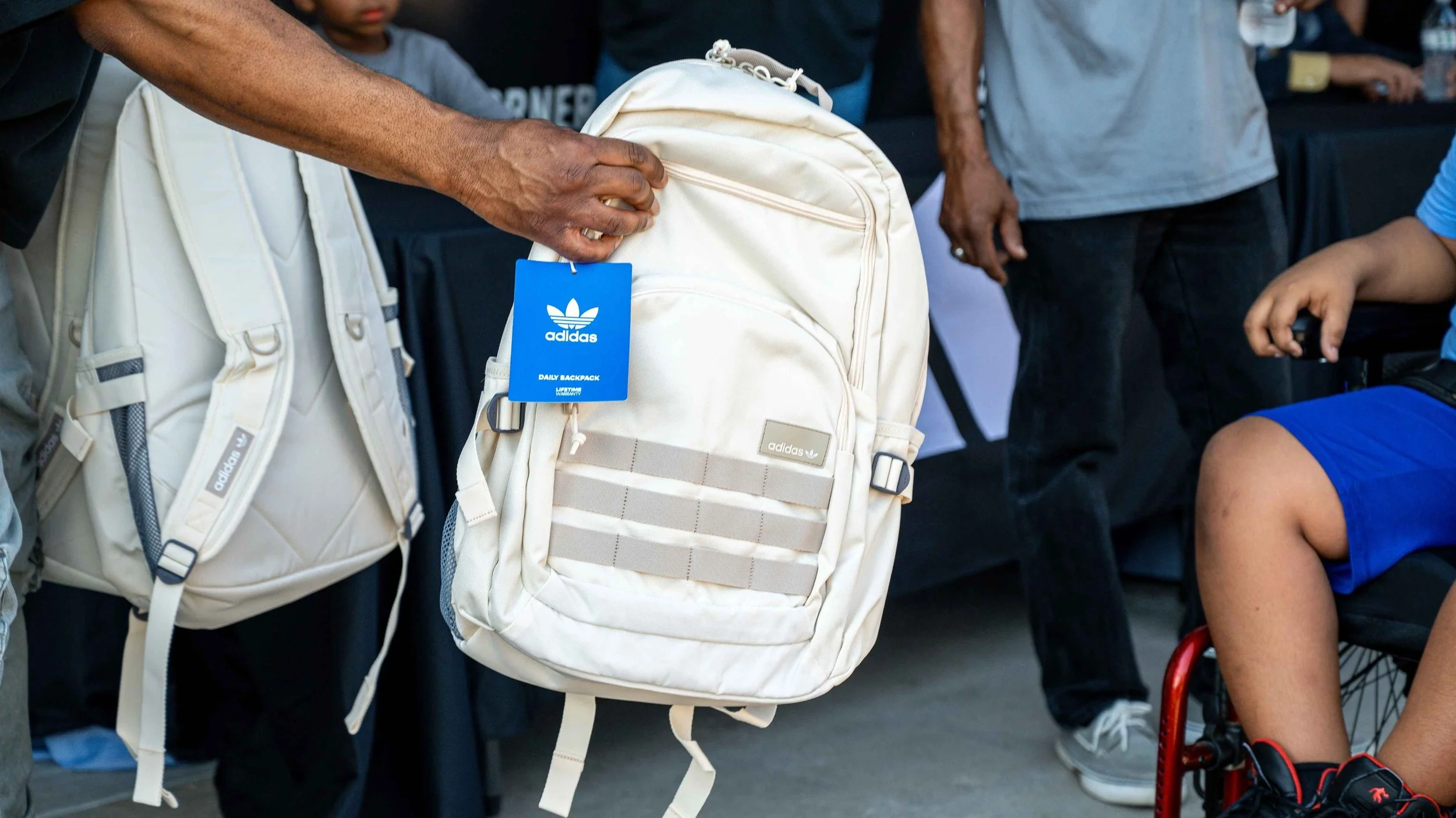 Person handing a white Adidas backpack to a child sitting in a wheelchair in a crowded indoor setting.