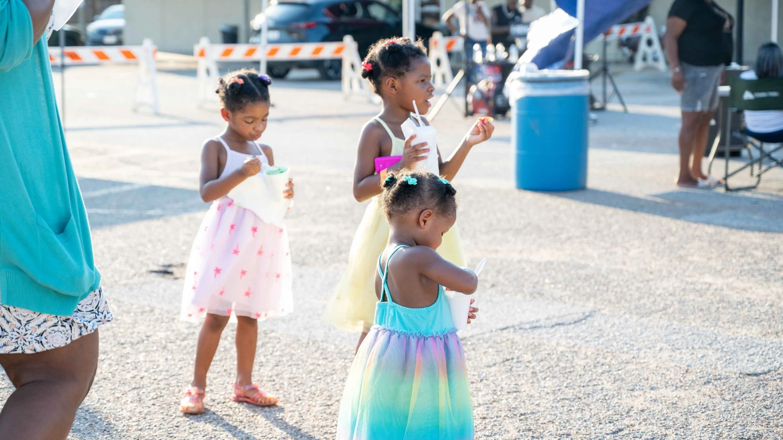 Three young girls in colorful dresses eating ice cream cones outdoors at a fair or festival, with adults and booths in the background.
