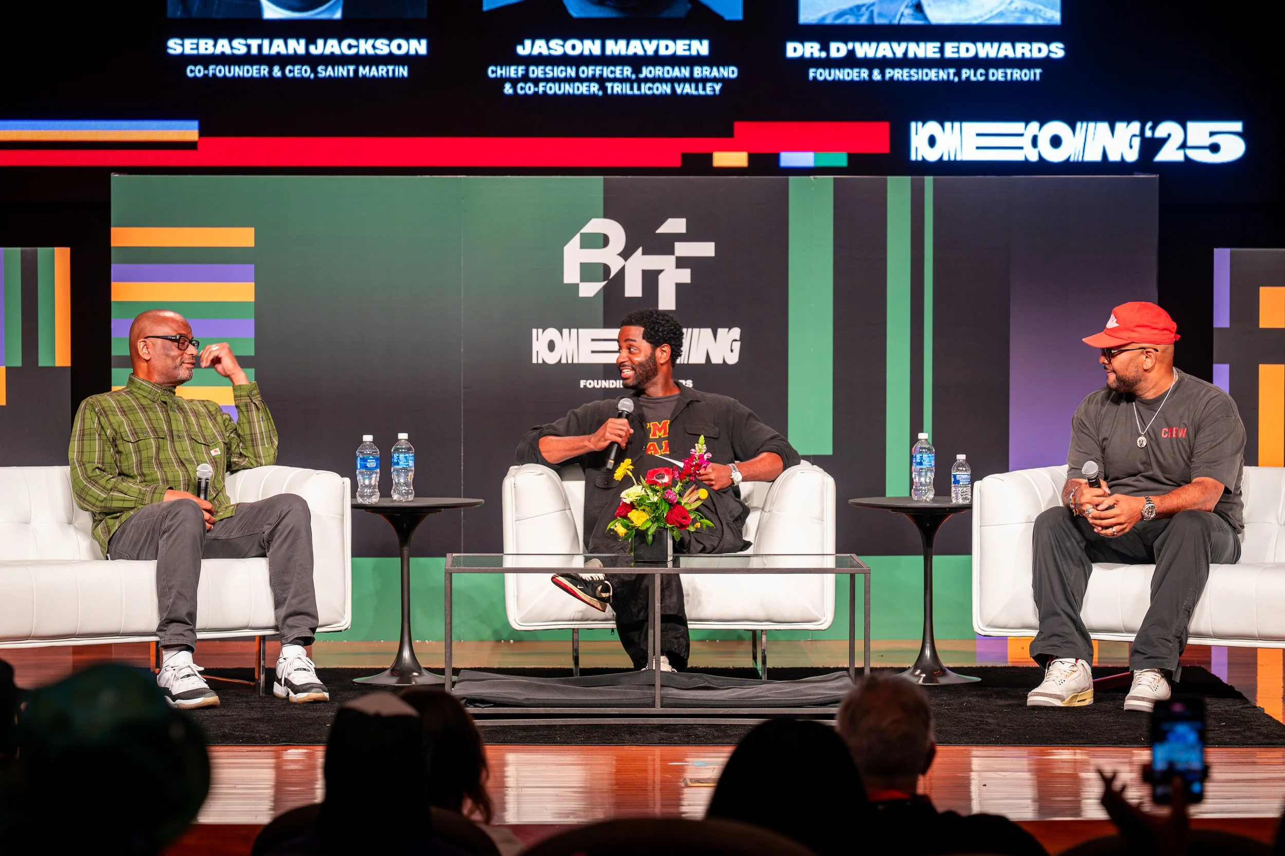 Three men are seated on stage in white chairs during a panel discussion, with a black backdrop featuring logos, a large green and black display screen, and three water bottles on small black tables. The man on the left wears a green plaid shirt, the man in the middle wears a black jacket and t-shirt and is holding a microphone, and the man on the right wears a red cap and a gray shirt. There are audience members visible in the foreground.