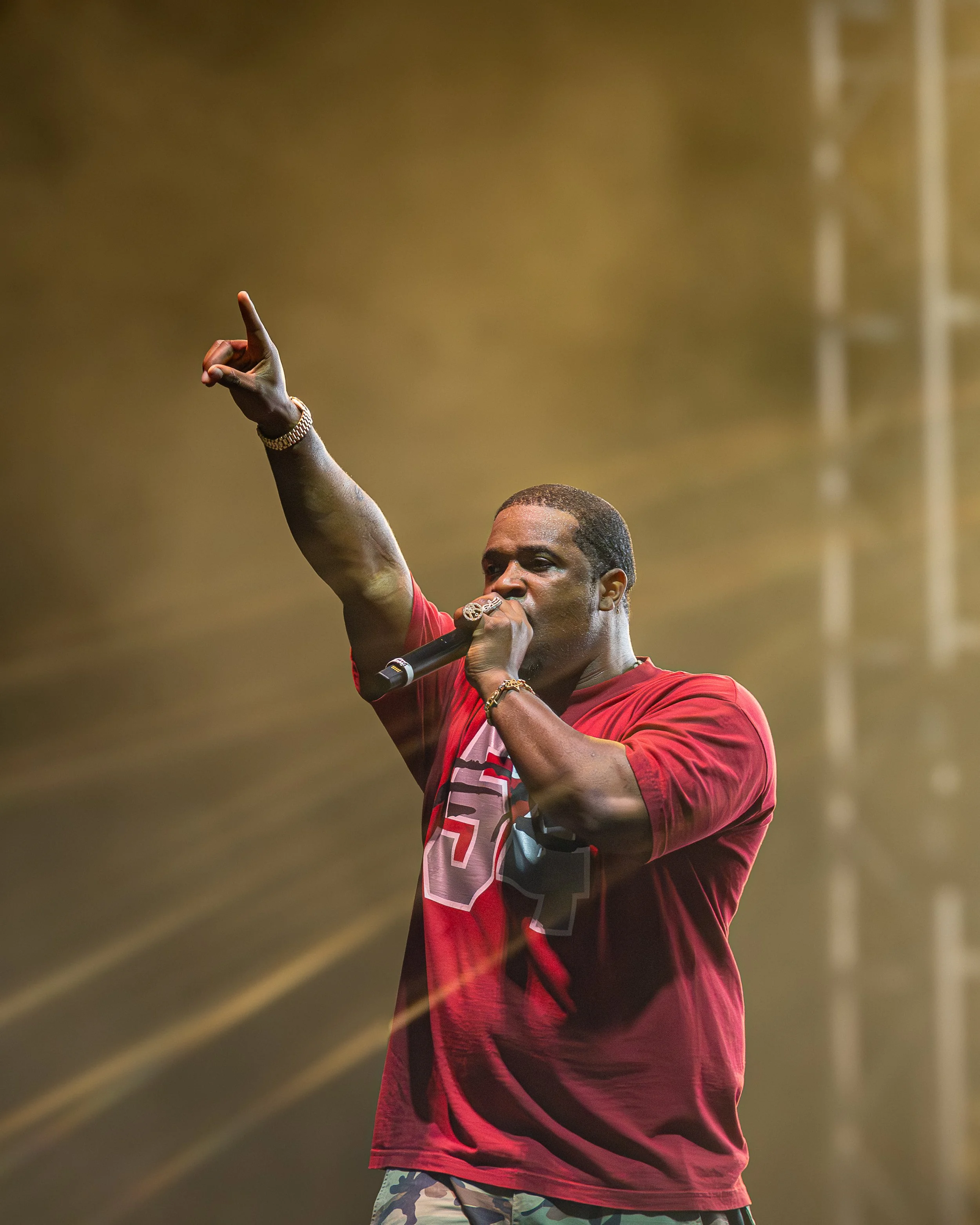 Male rapper performing on stage, holding a microphone in his right hand, wearing a red t-shirt, gold bracelets, and camouflage shorts, with stage lights in the background.