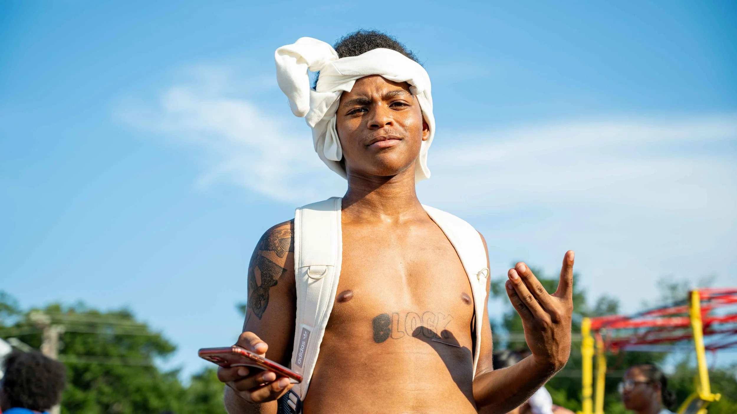 A shirtless young man with a towel wrapped around his head, holding a phone in one hand and making a peace sign with the other, outdoors on a sunny day.