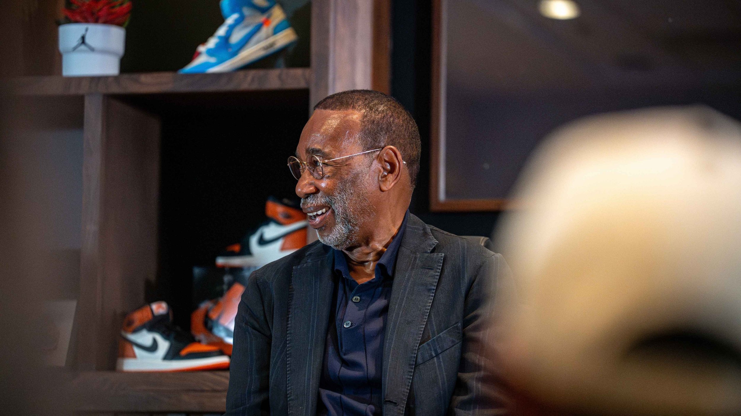 An older man with glasses and a beard, smiling and talking, dressed in a dark pinstripe blazer and dark shirt, sitting indoors near a shelf with sneakers and decorative items.