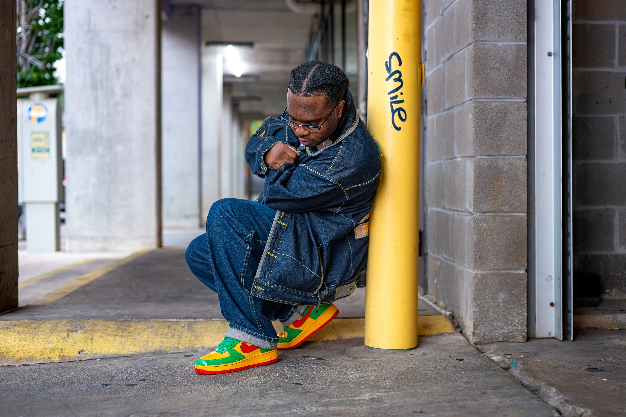 A man with glasses and braided hair wearing a denim jacket and jeans, colorful sneakers, sits on the ground outside a building, leaning against a yellow pole