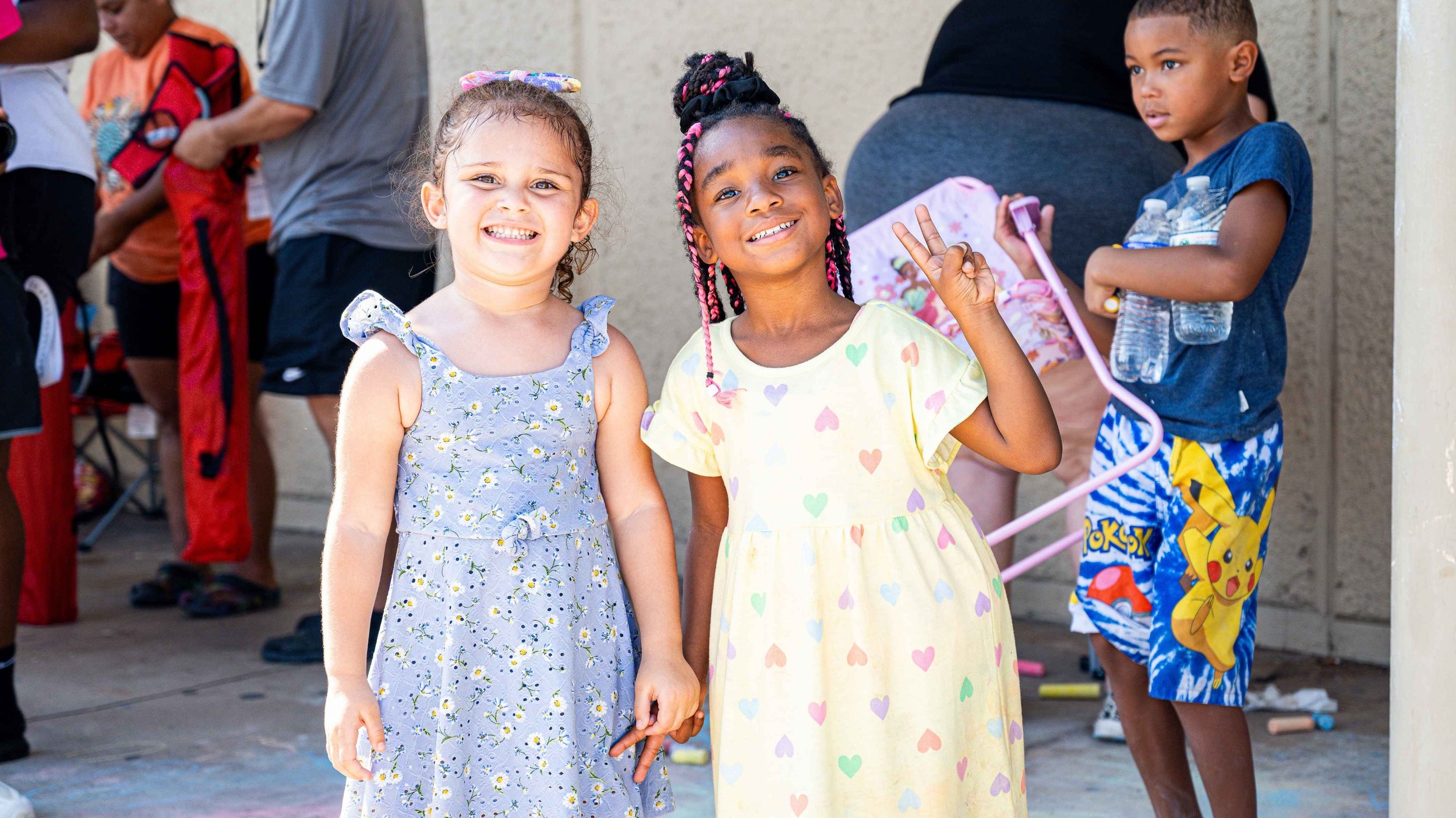 Two young girls smiling and holding hands, one wearing a blue floral dress and the other wearing a yellow dress with colorful hearts, standing outside at a busy event with children and adults in the background.