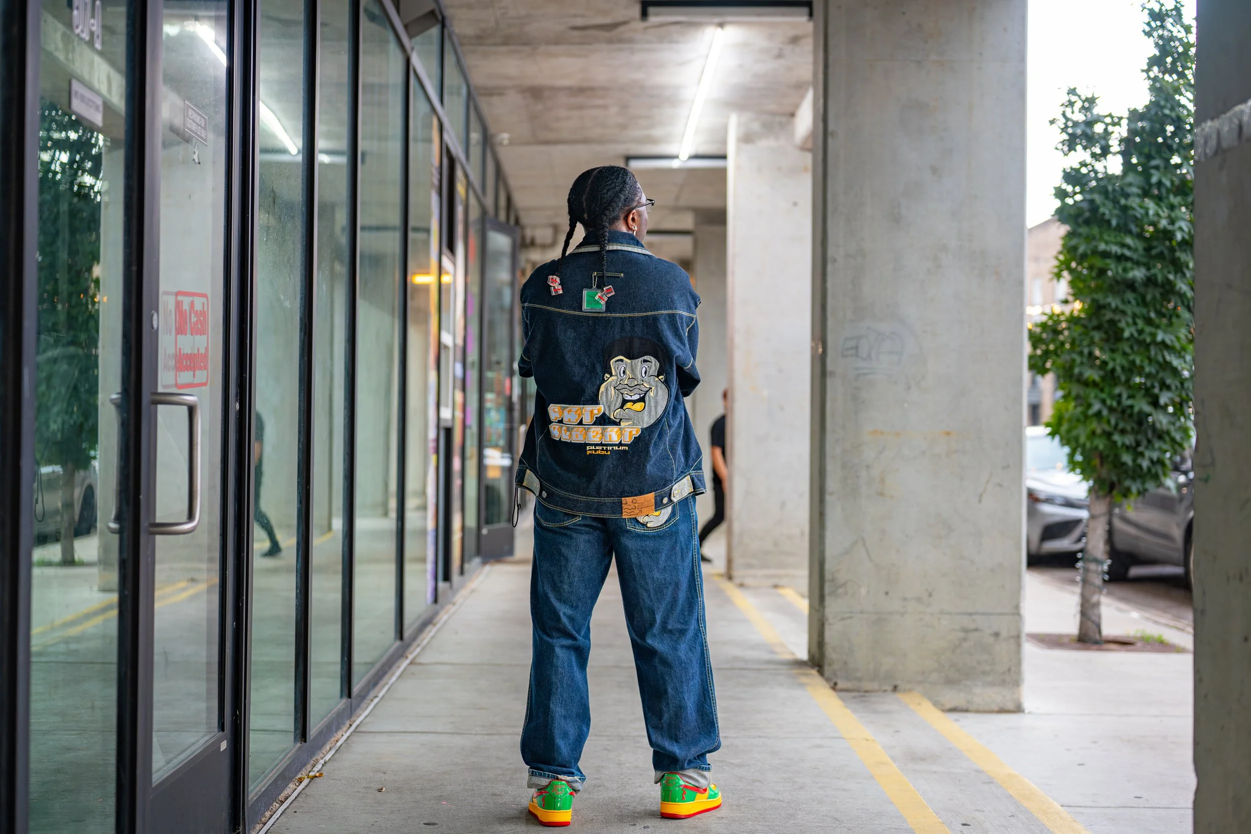 A person standing outside a store with glass doors, wearing a dark denim jacket with patches and designs on the back, loose jeans, and colorful sneakers, facing away.
