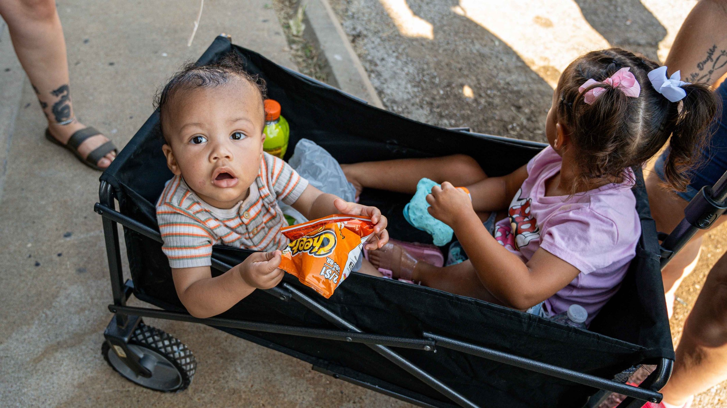 A young boy sitting in a stroller holding a bag of Cheetos with a surprised expression, next to a girl with pigtails holding a blue toy.