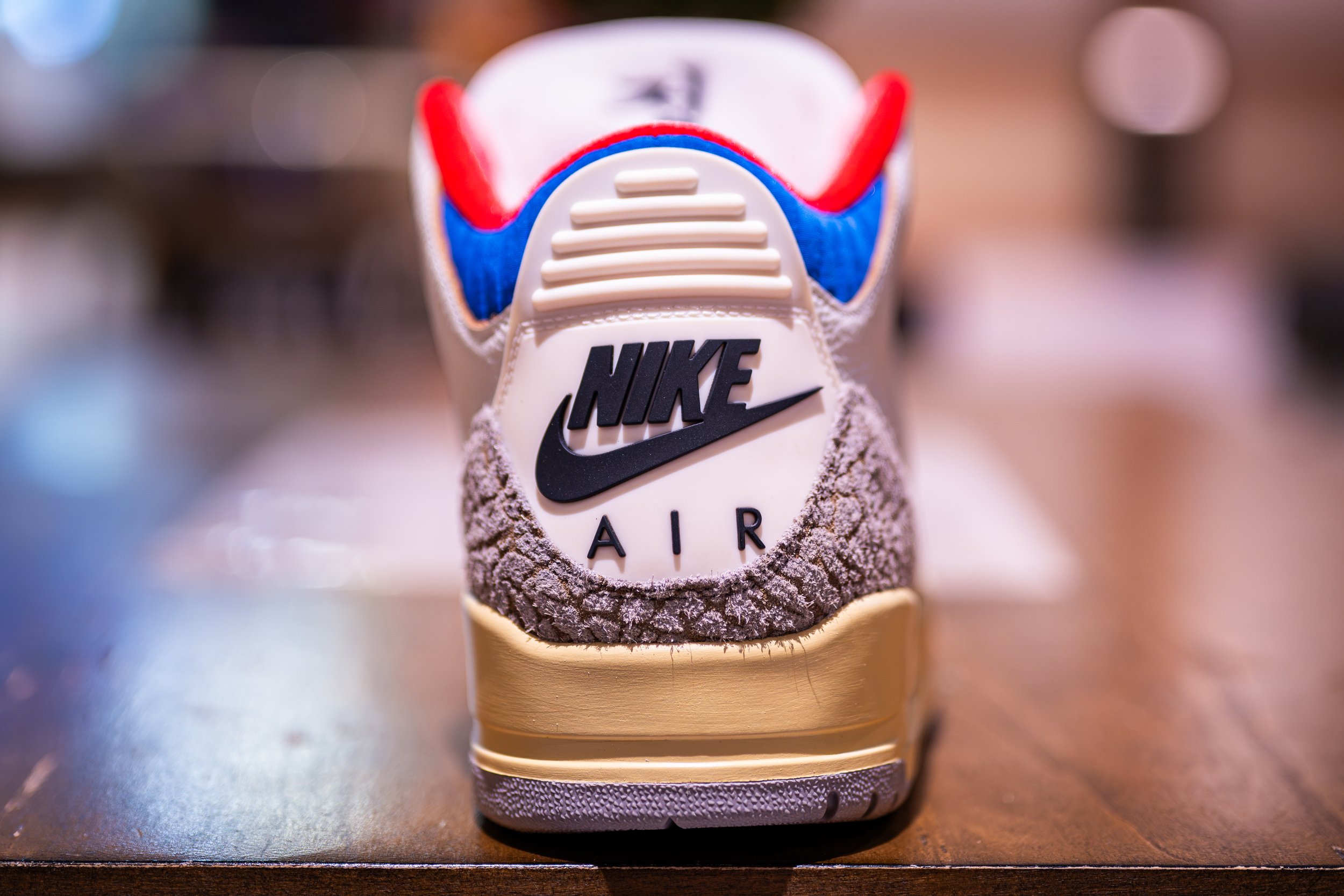 Close-up of the back of a Nike Air sneaker with a prominent Nike logo, made of a mix of beige, brown, white, red, and blue materials, sitting on a wooden surface.