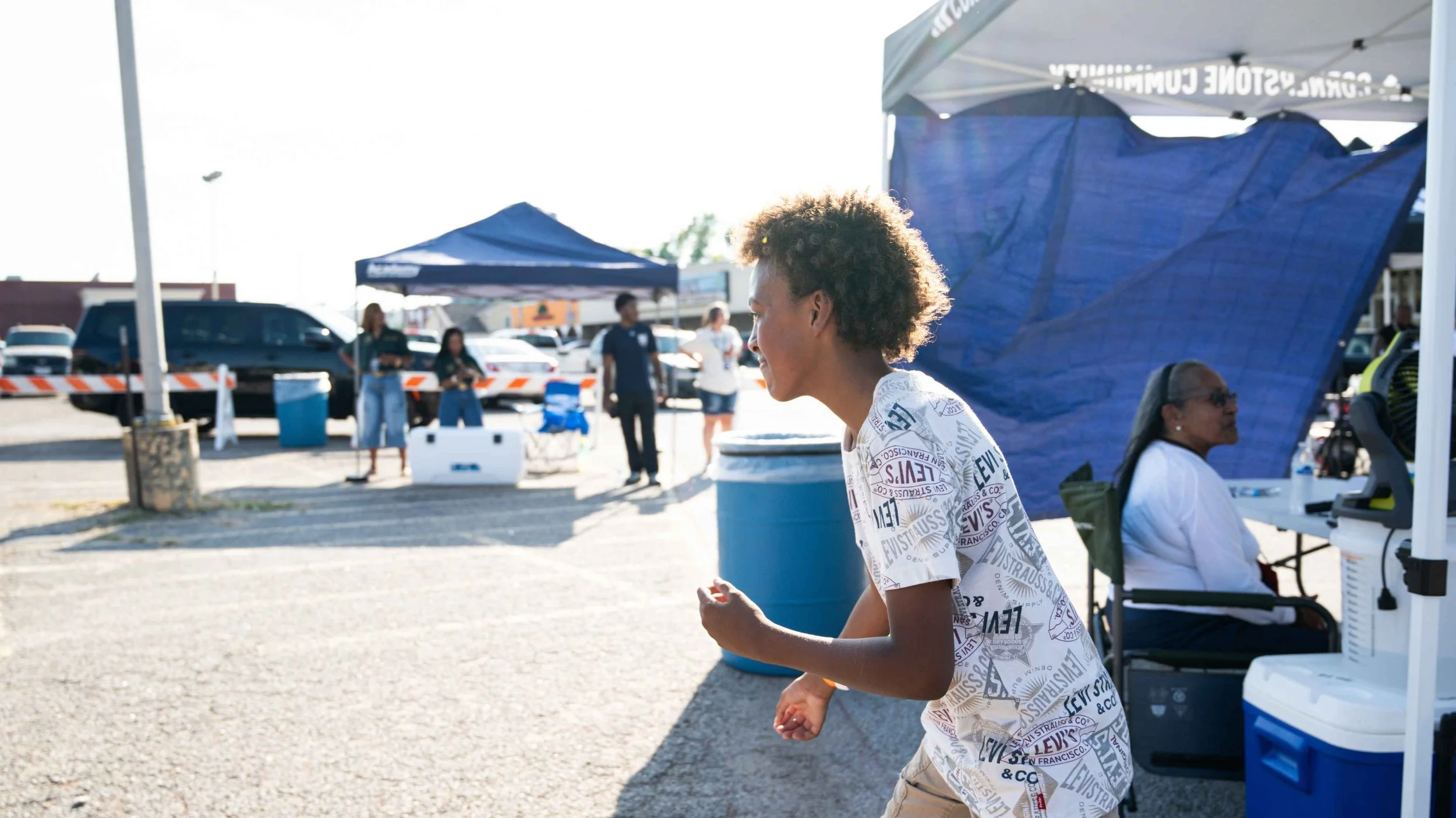 A young boy with curly hair in a white graphic t-shirt laughing and playing outside at a community event or festival, with tents and other people in the background.