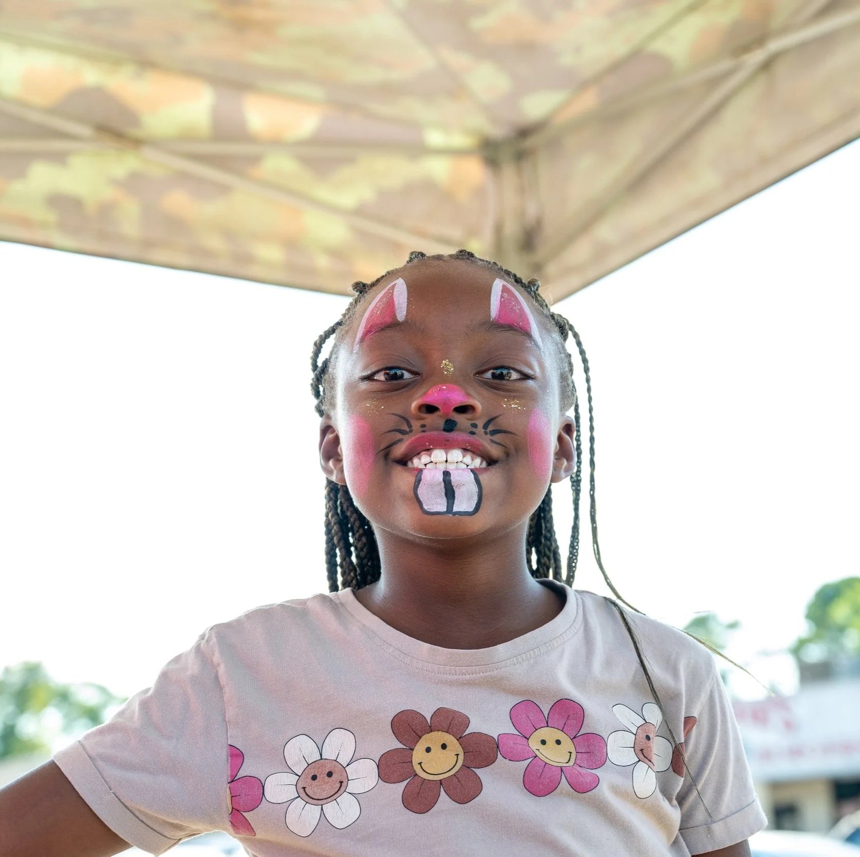 A smiling young girl with face paint resembling a bunny, wearing a T-shirt with smiling flowers, standing outdoors under a tent.