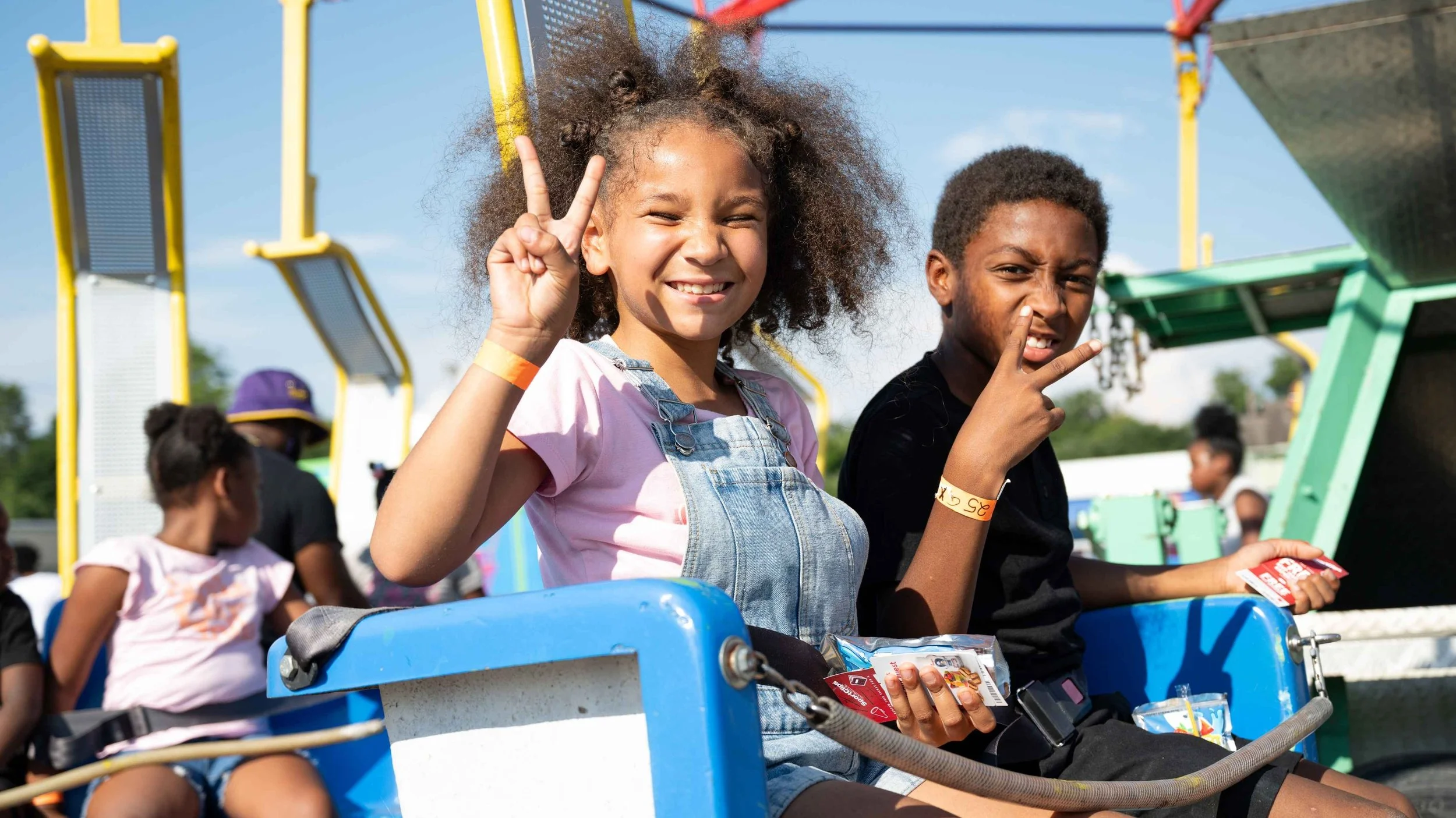 Two children sitting on a carnival ride, making peace signs with their hands, smiling, with snow cone treats in their hands, and other children and amusement park equipment in the background.