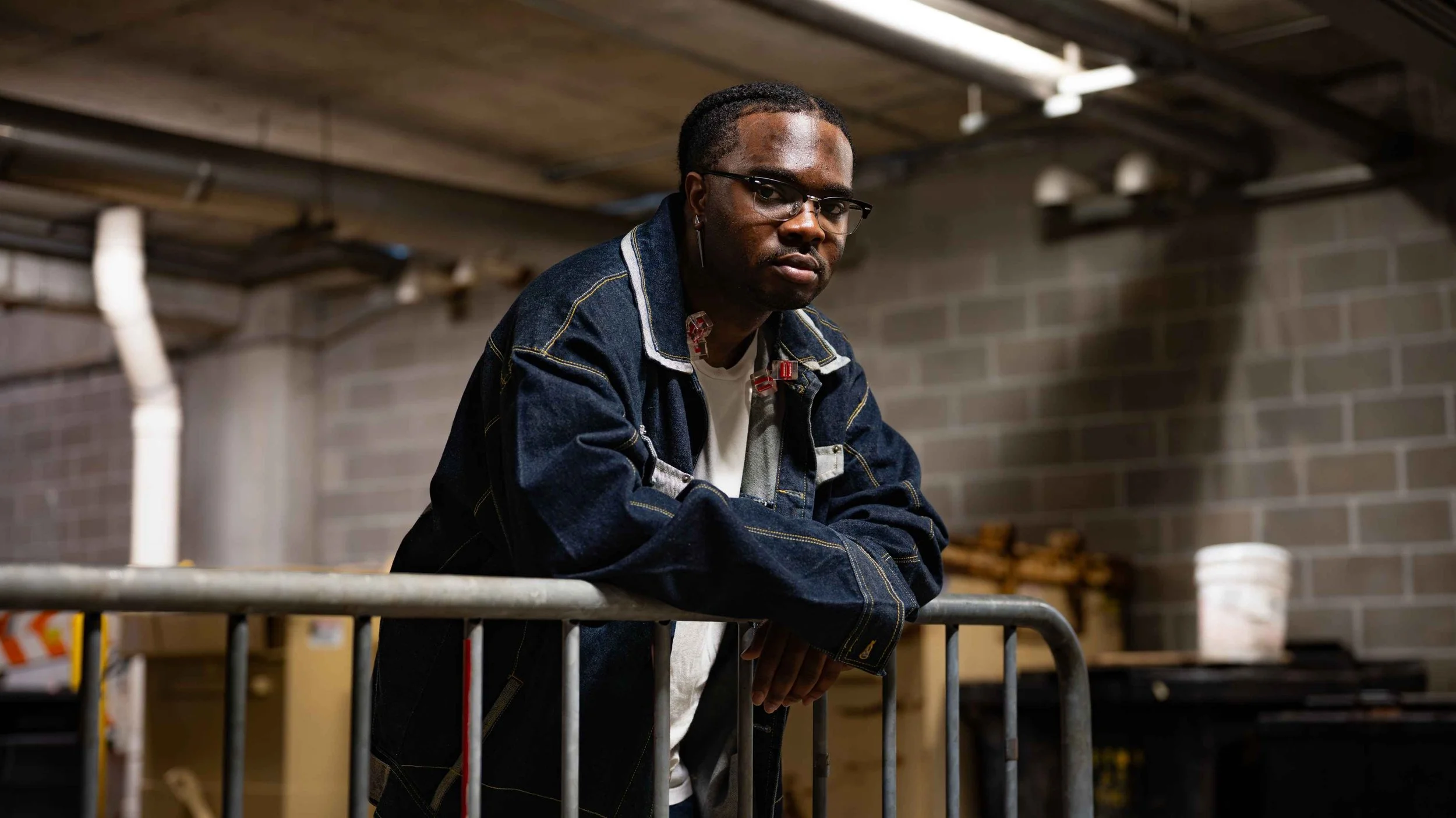 A man with glasses and earrings leaning on a metal railing in an industrial or storage room with brick walls and pipes.