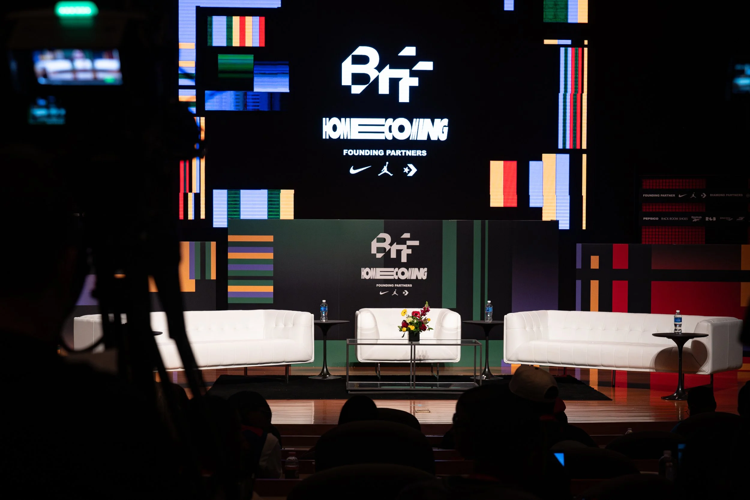 Stage setup for an event with three white sofas, a table with a flower arrangement, and a backdrop displaying the logo for 'BE HOME COMING' with sponsor logos, and a large digital screen with colorful graphics.
