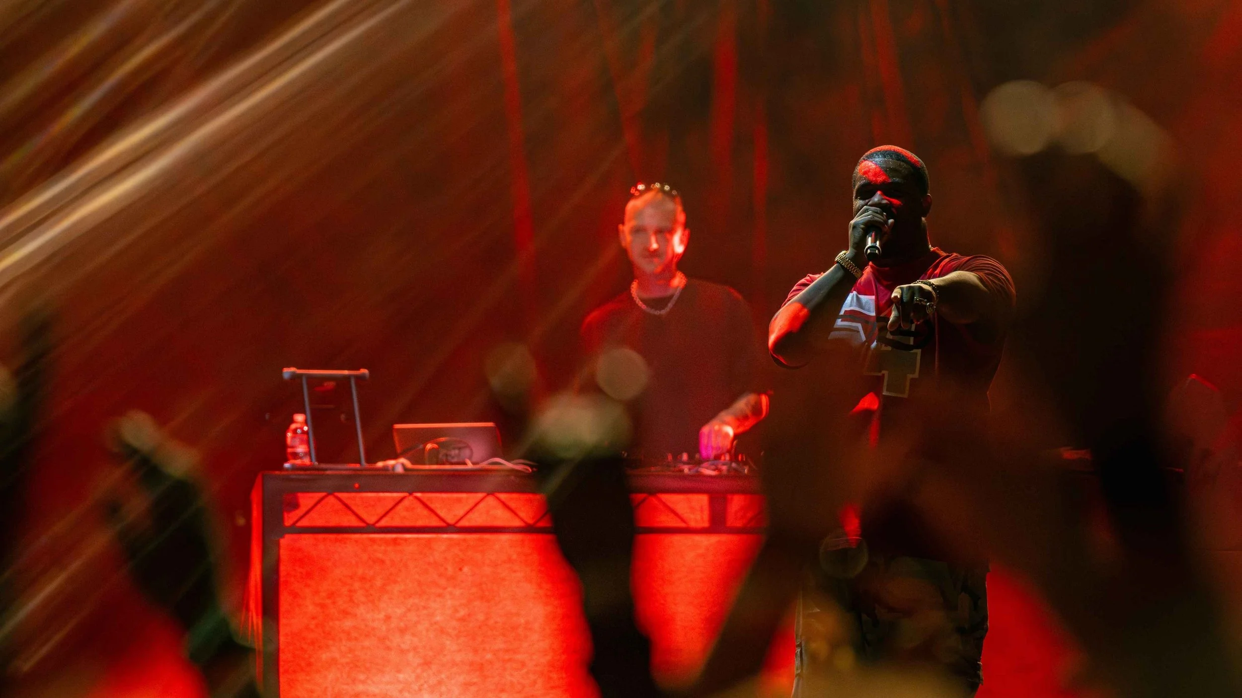 Two performers on stage with DJ equipment, one holding a microphone and pointing towards the camera, in a dimly lit environment with red and orange lighting.