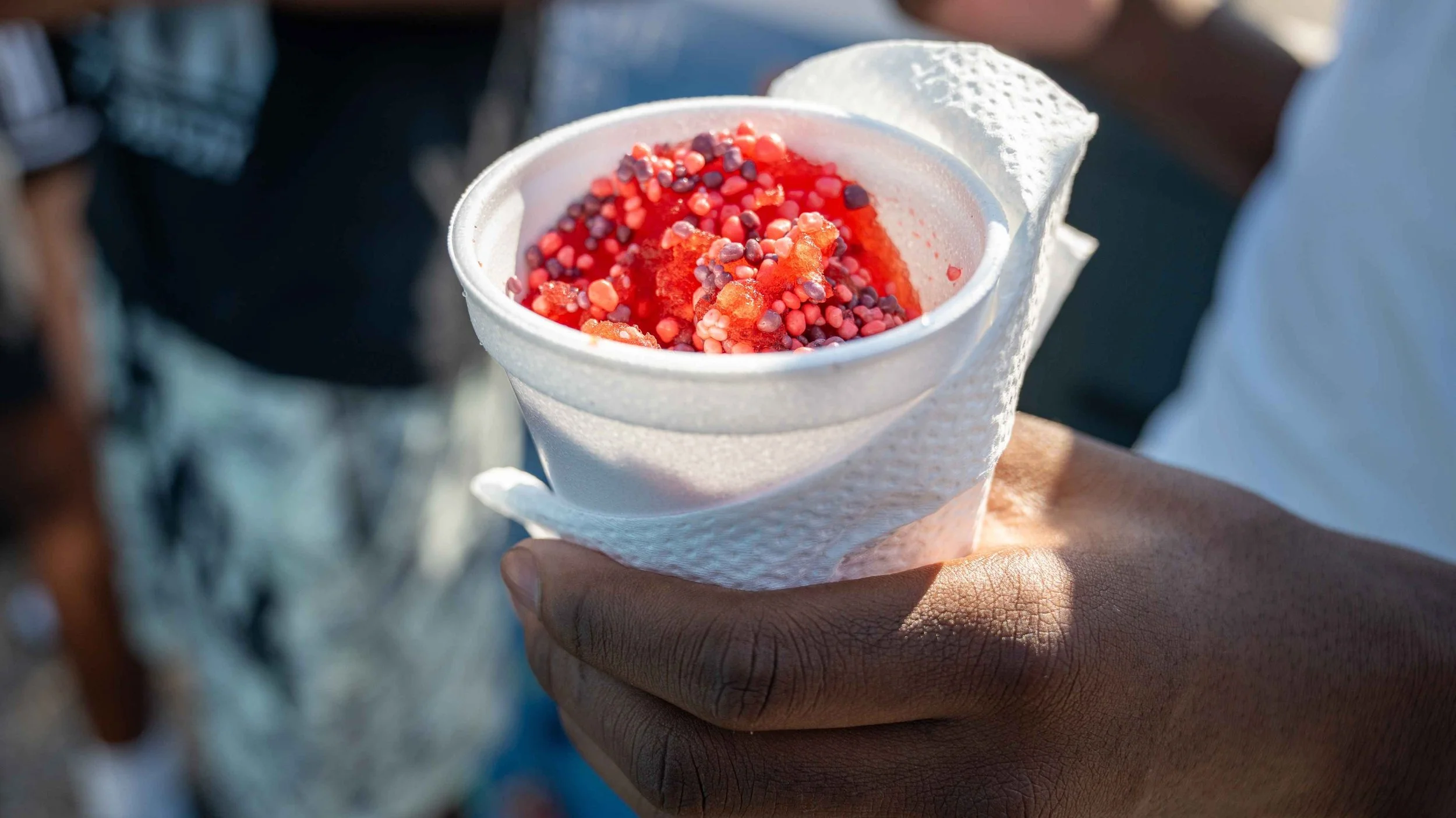 Close-up of a hand holding a cup of frozen treat topped with red and purple sprinkles, wrapped in a white napkin.