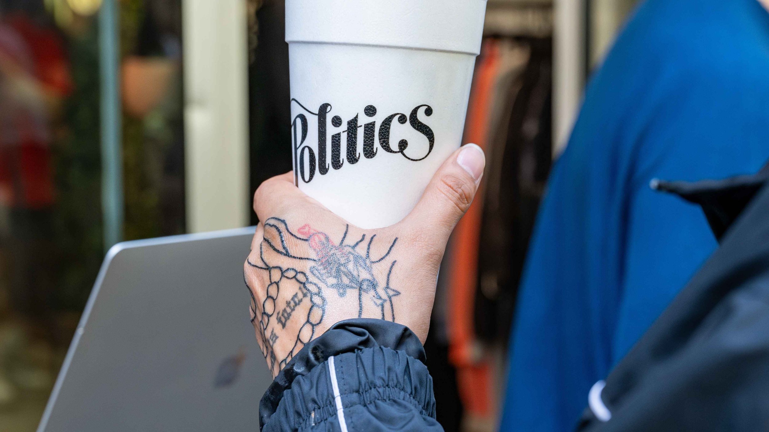 A person holding a white cup with the word 'Politics' written on it. The person has tattoos on their hand and is wearing a dark jacket. Part of a laptop is visible in the background.