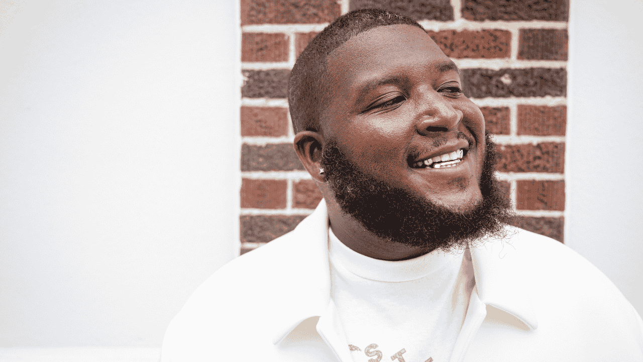 A smiling man with a beard and short hair, wearing a white jacket and shirt, standing in front of a brick wall.