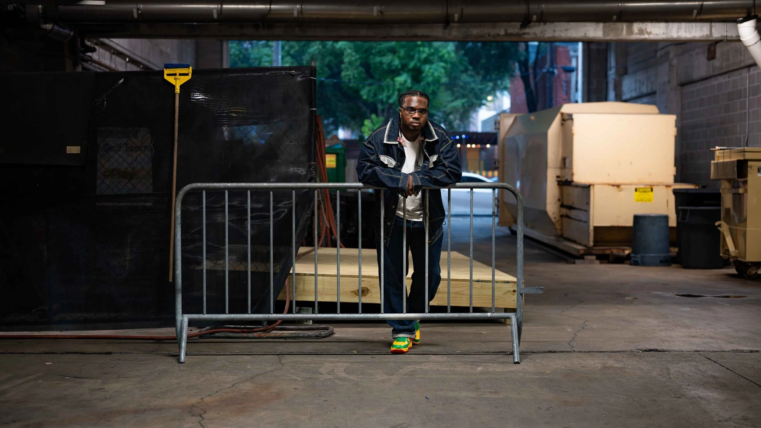 A man in a dark jacket and colorful sneakers stands with his arms on a metal barricade in an urban alley, with construction and utility equipment in the background.