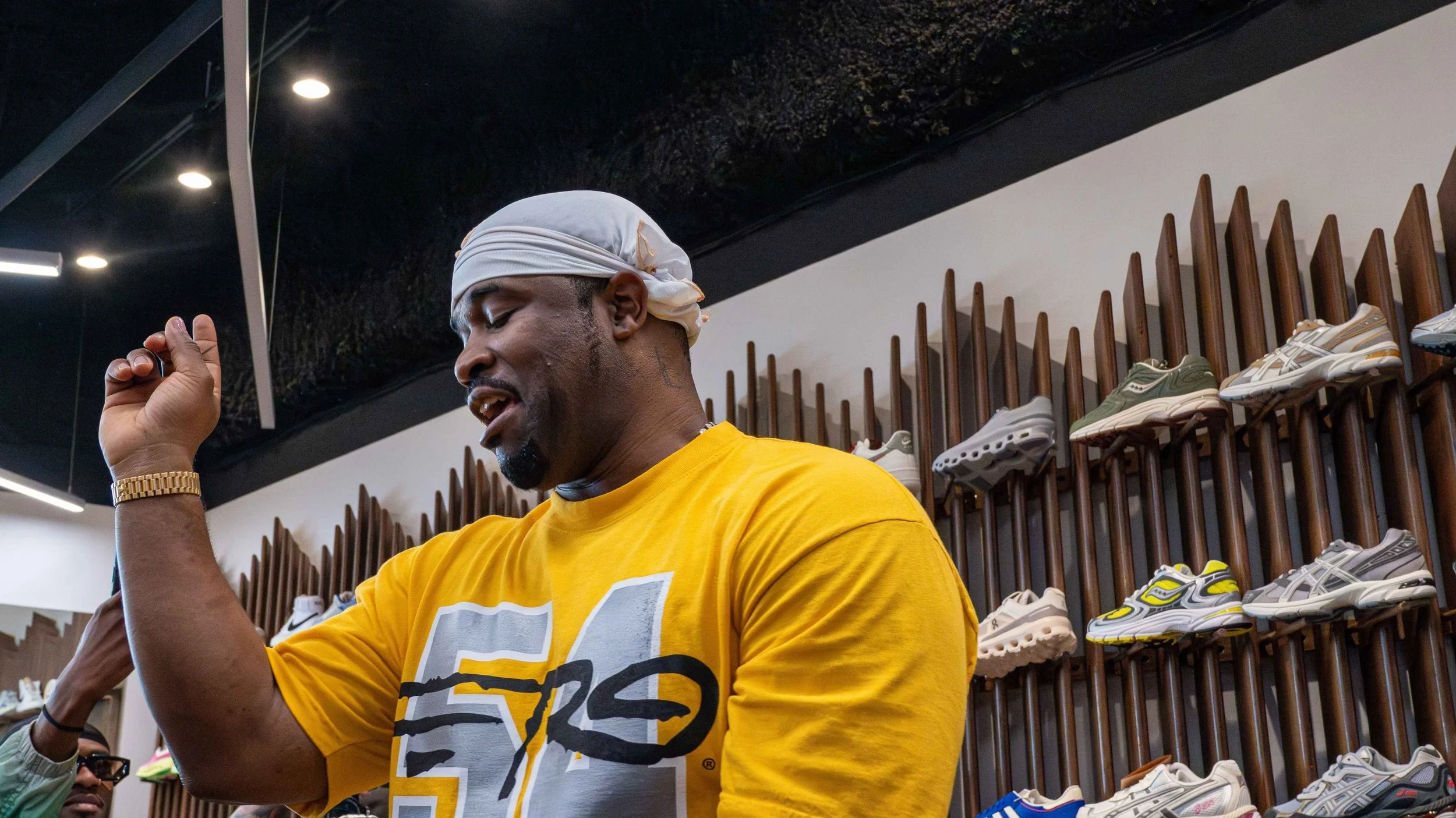 A man wearing a yellow T-shirt and a white headscarf, with a gold watch, standing inside a shoe store, with shelves of sneakers behind him.