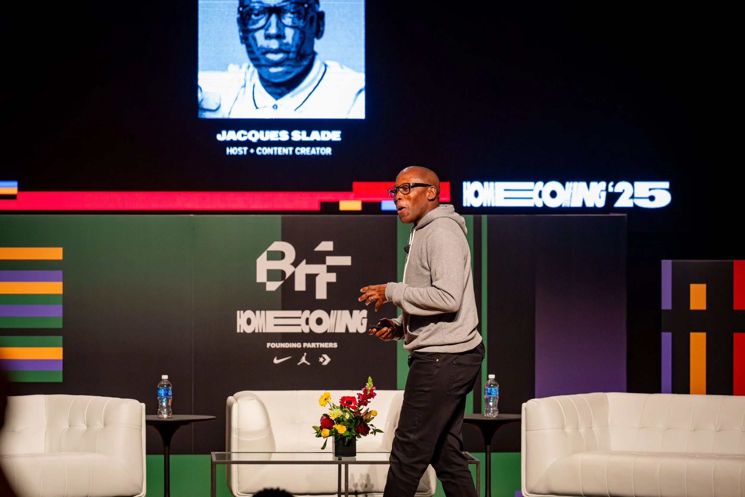 A man in glasses and a gray hoodie speaking on stage at the Homecoming '25 event, with a slide showing Jacques Slade, host and content creator, projected behind him, alongside colorful striped and block patterns.