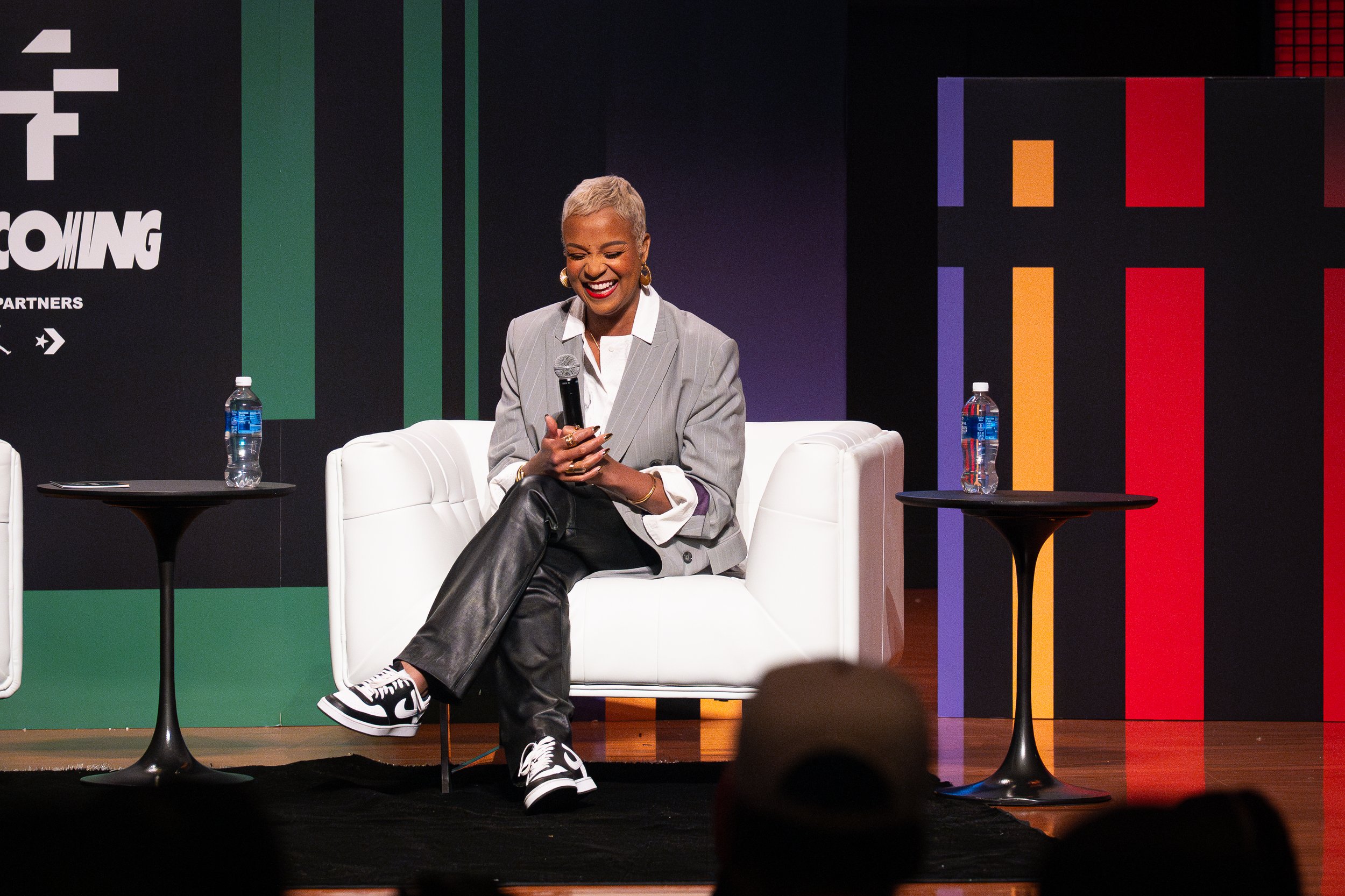 A woman sitting on a white couch, smiling and holding a microphone on stage with a colorful background and two small tables with water bottles.