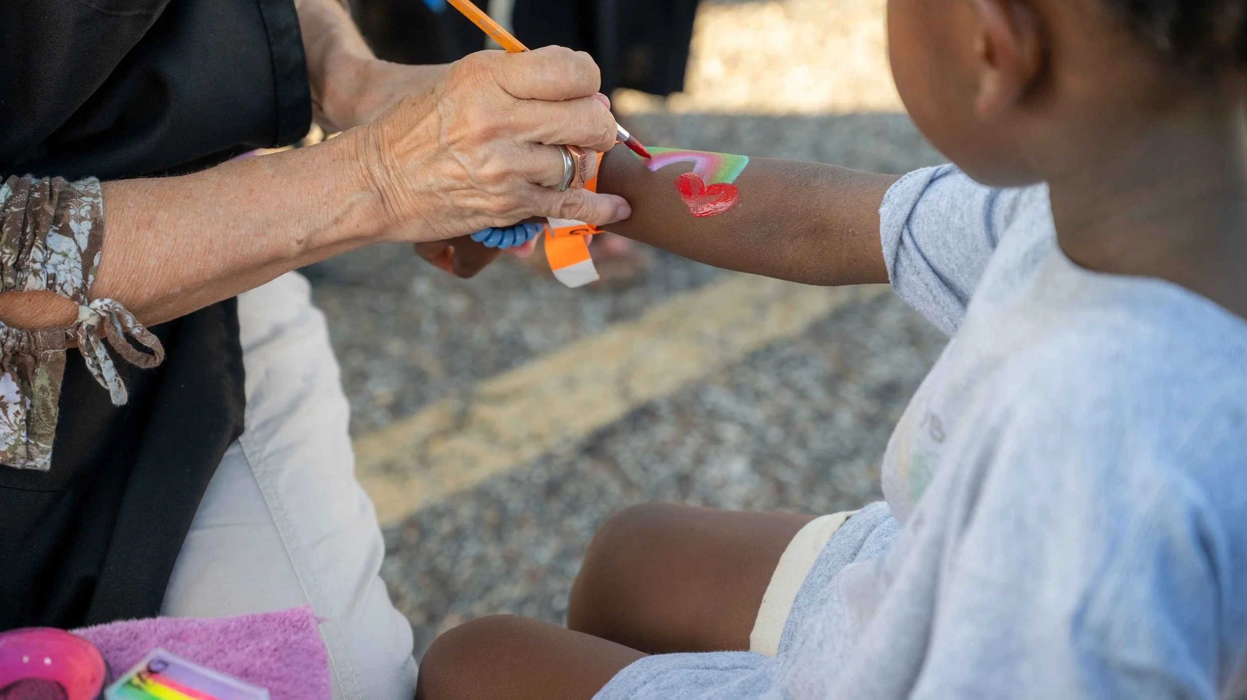 An older person is painting a colorful rainbow with a red heart on a young child's outstretched arm during a daytime outdoor activity.