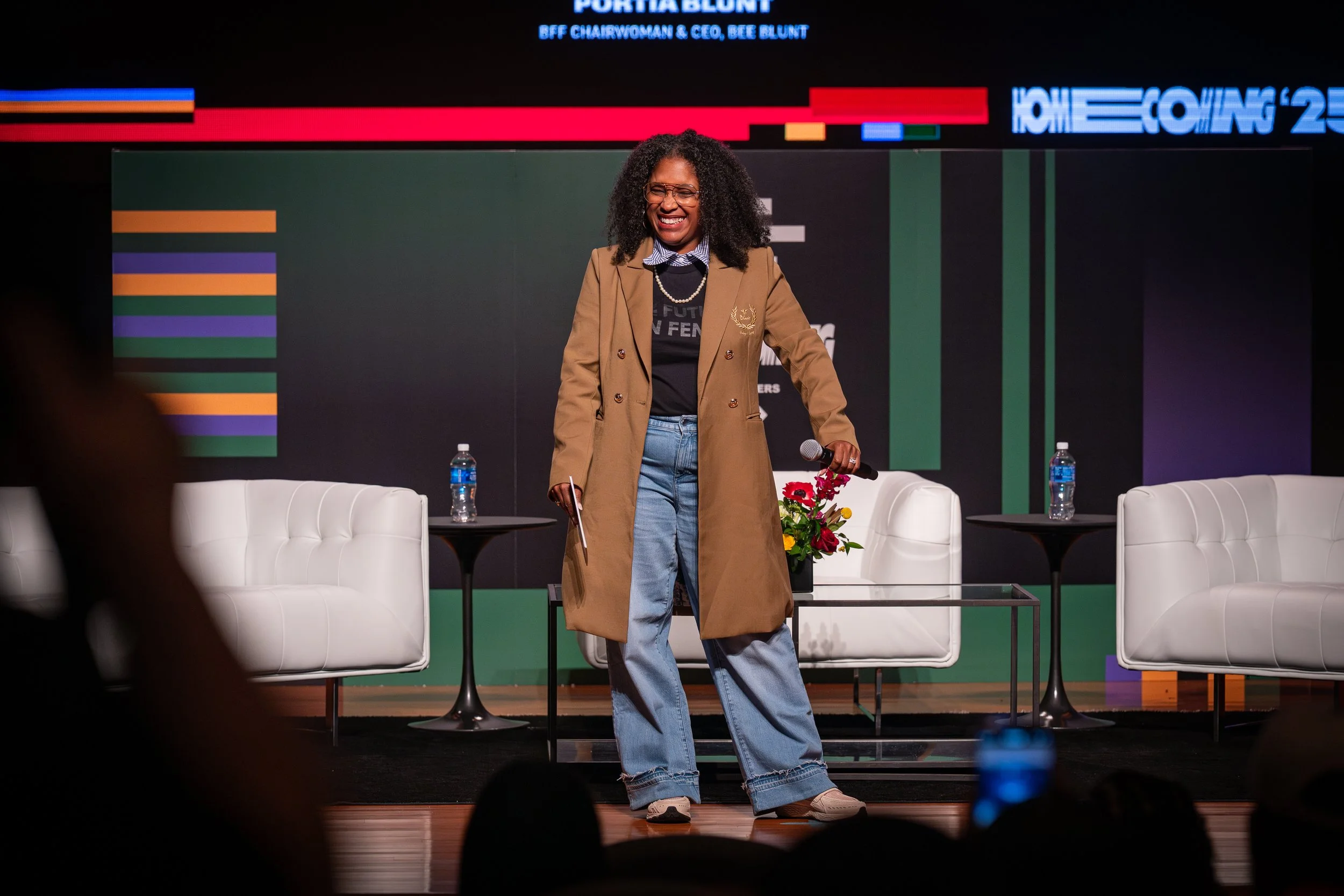 A woman with curly black hair, glasses, and a bright smile standing on a stage with a microphone in her right hand. She is wearing a tan trench coat, black T-shirt, blue jeans, and beige sneakers. Behind her are white chairs, small tables with water bottles, and a flower arrangement. The background features colorful lines and text indicating a conference or event.