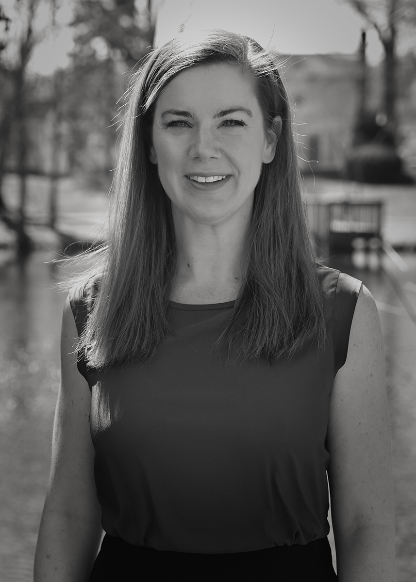 Black and white photo of a smiling woman with shoulder-length hair outdoors, with a blurred background of trees and a pond.