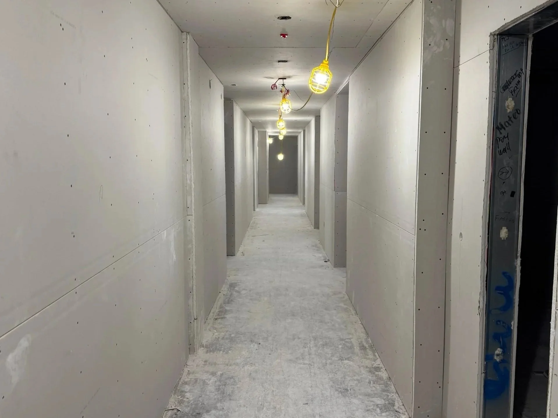 A long, white-walled hallway under construction with exposed light bulbs hanging from yellow cords, partially finished doorways, and a concrete floor.