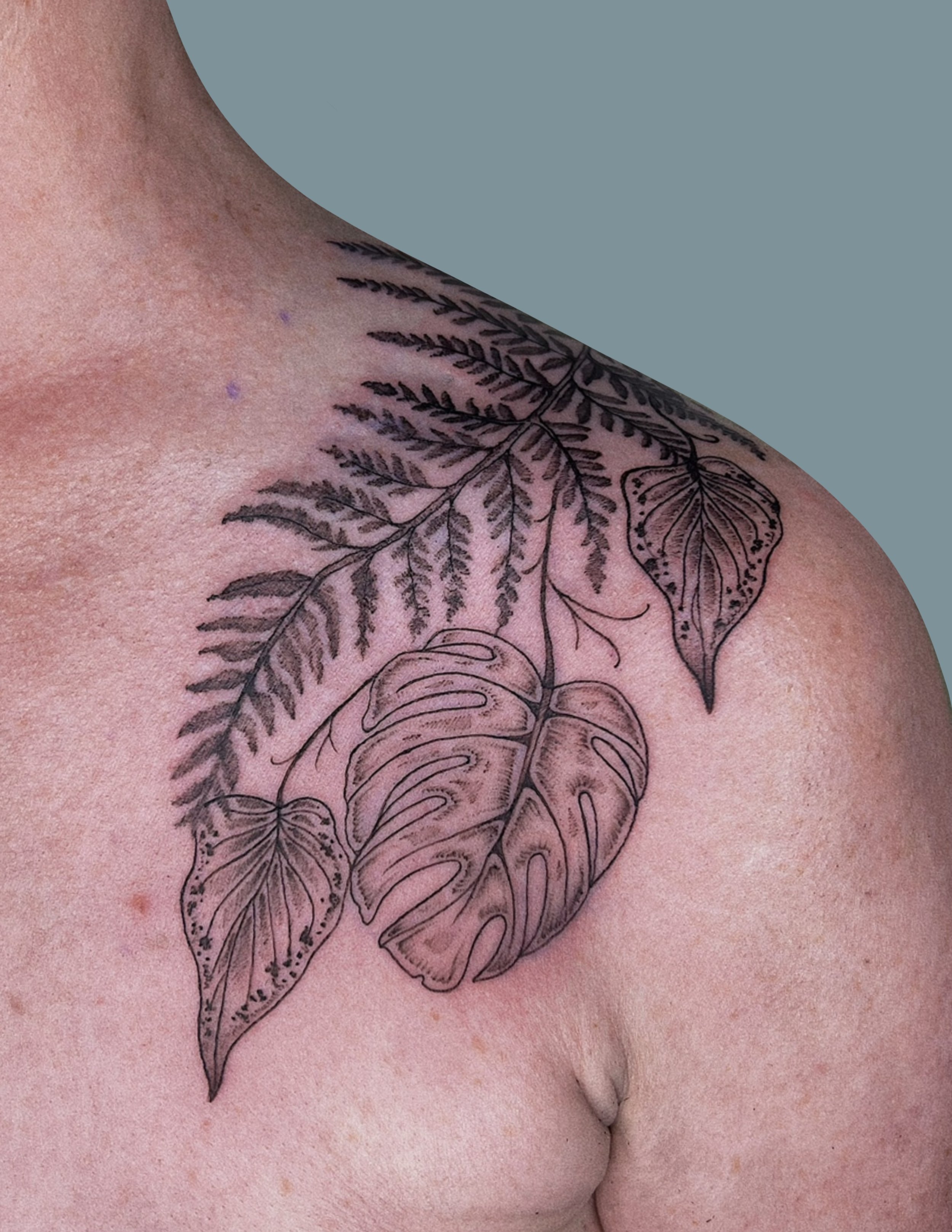 Tattoo of tropical leaves, including monstera and palm fronds, on someone's shoulder.