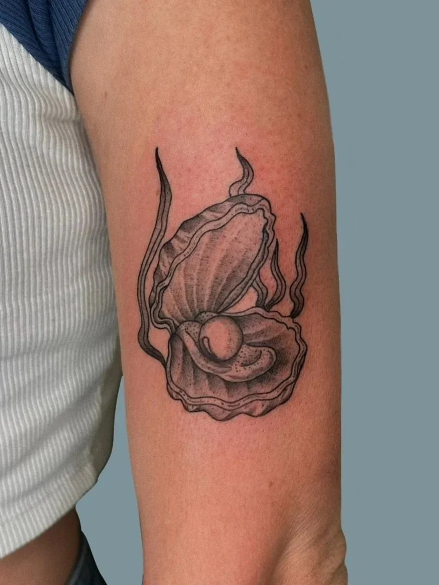 Tattoo of a clam shell with a pearl inside, drawn with black ink and shading, on a person's upper arm.
