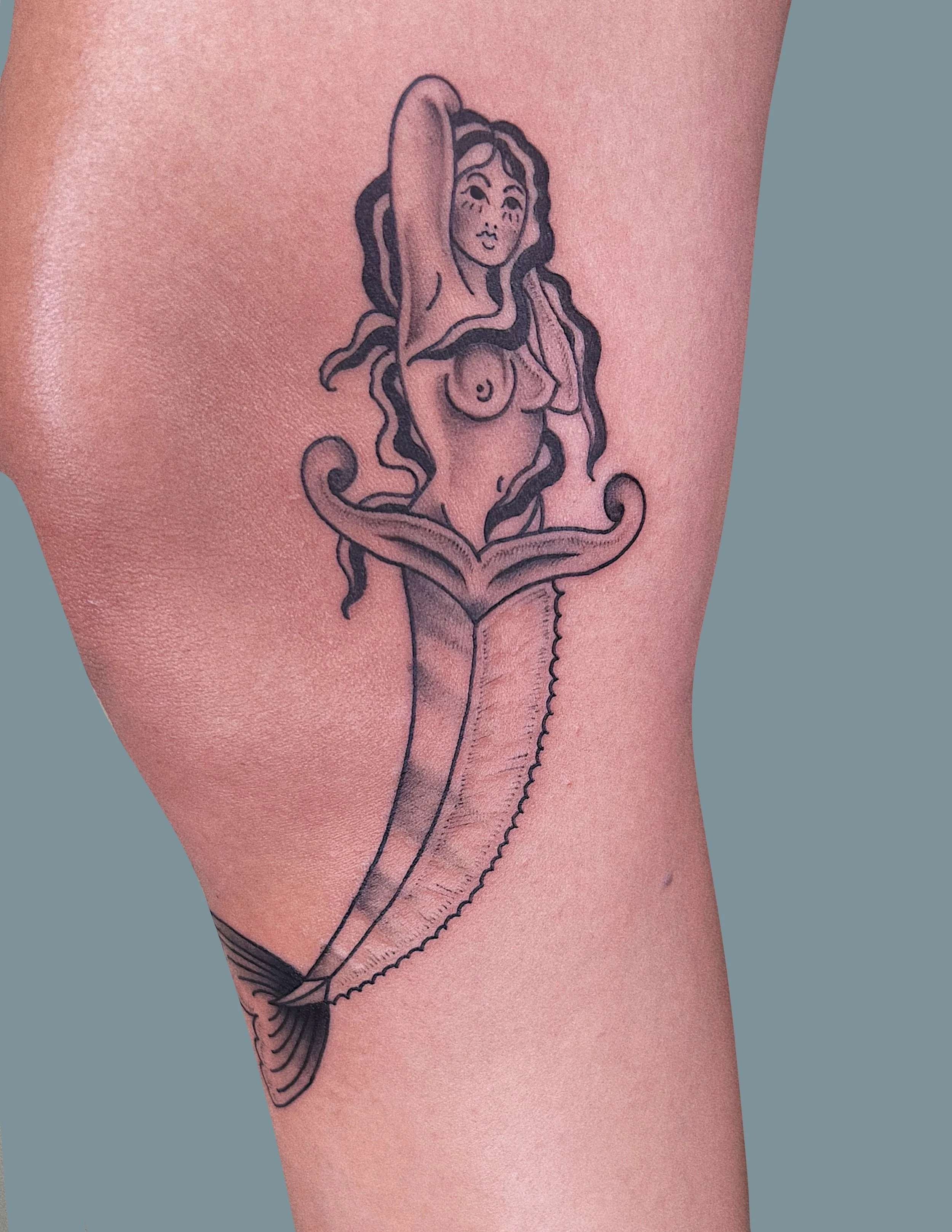 Tattoo of a mermaid with long, curly hair, and a calm face, emerging from a large, ornate seashell on person's upper arm.