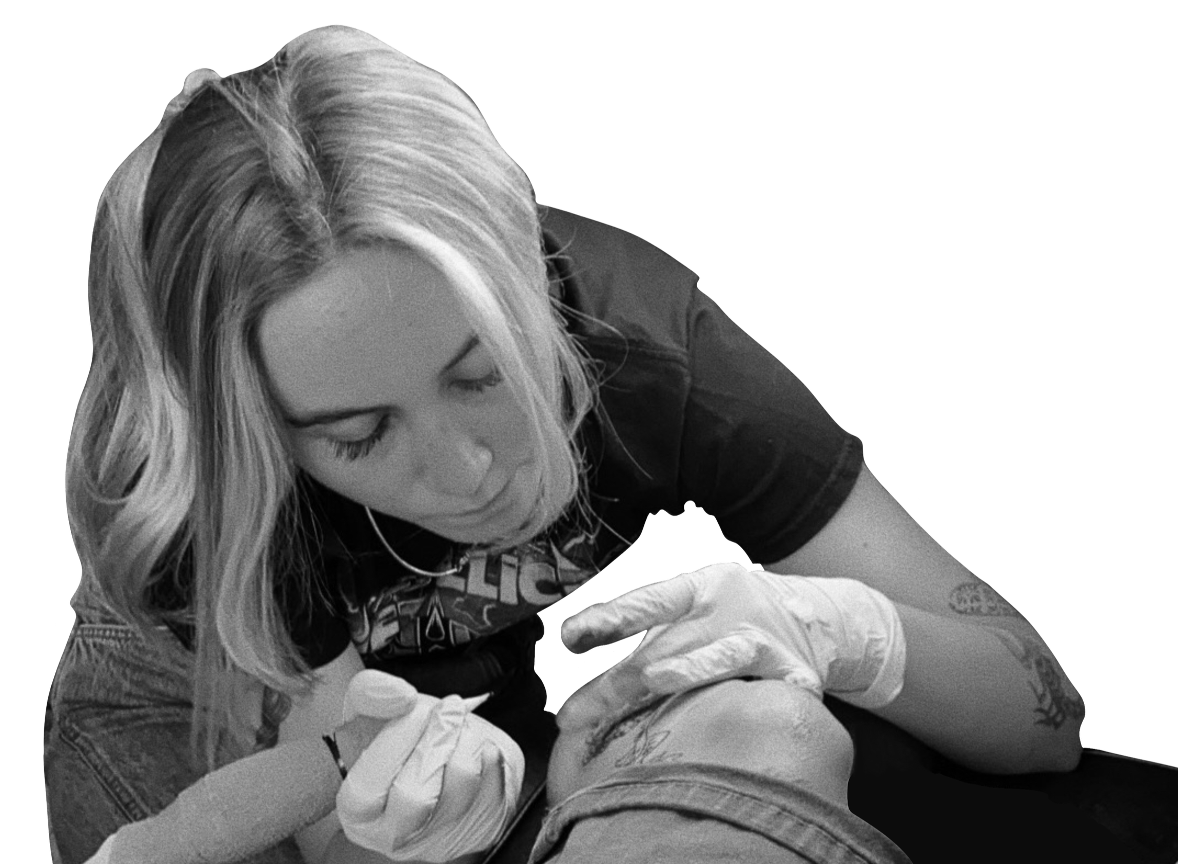 A woman with blonde hair, wearing a black t-shirt and white gloves, is tattooing a person's leg, with her head tilted downward, focused on the tattooing process.