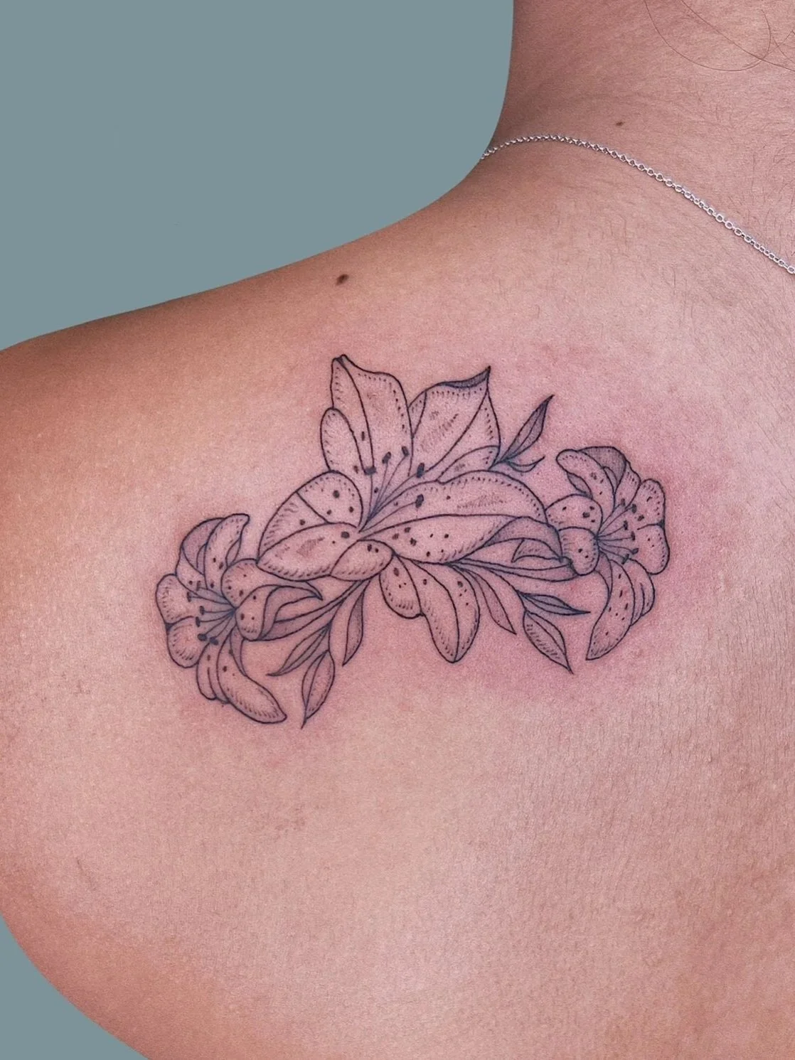 Close-up of a person's shoulder with a floral tattoo of lilies and leaves in black ink.