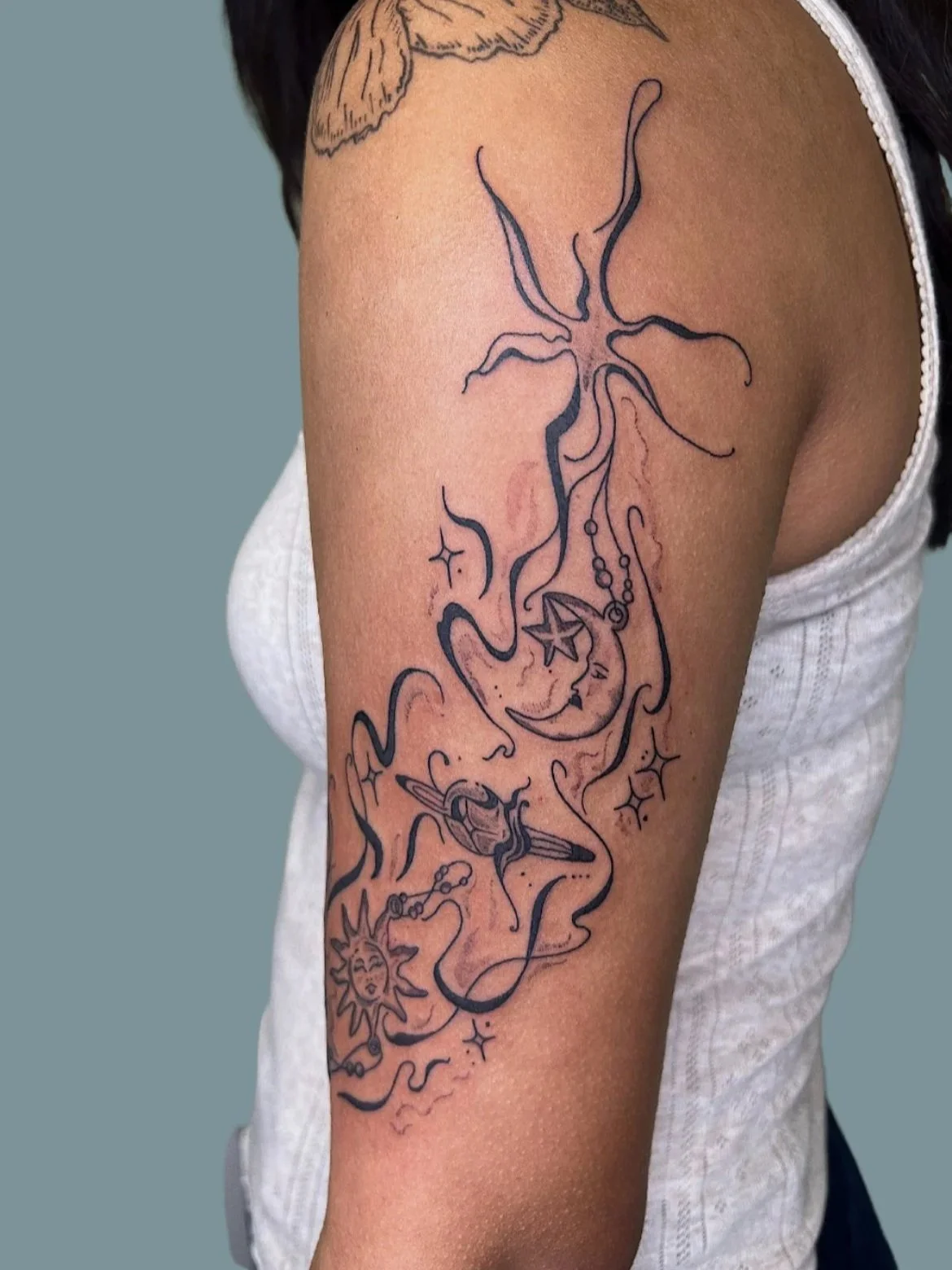 Tattoo of a moon with a face, surrounded by celestial and mystical symbols, including stars, a sun with a face, and abstract lines on a person's upper arm.