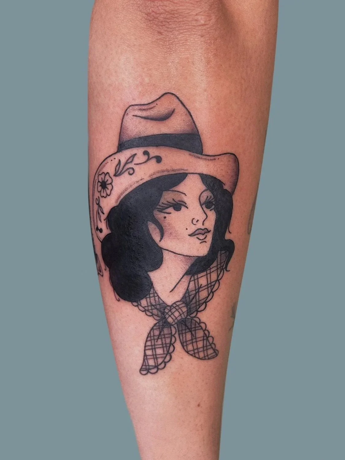 A tattoo of a woman with dark hair wearing a wide-brimmed hat with floral and vine details, and a lace neck scarf.