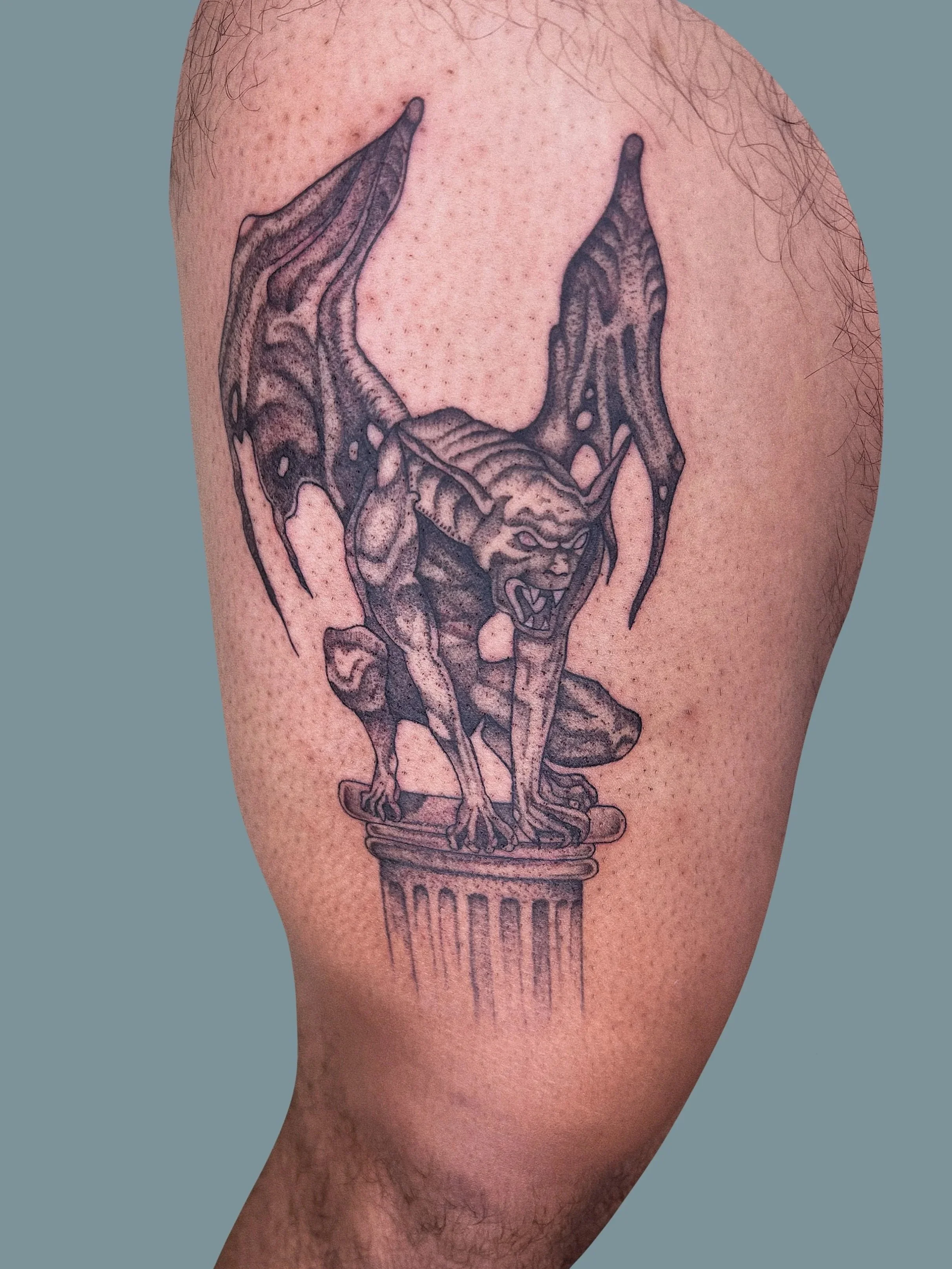 Tattoo of a bat-like creature with wings, crouching on top of a pedestal, with aggressive facial expression and sharp features.