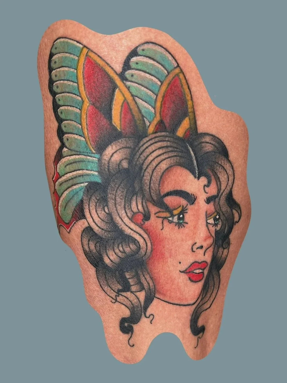 Tattoo of a woman with dark curly hair, colorful butterfly or winged headdress, and makeup with red lipstick and eye makeup.