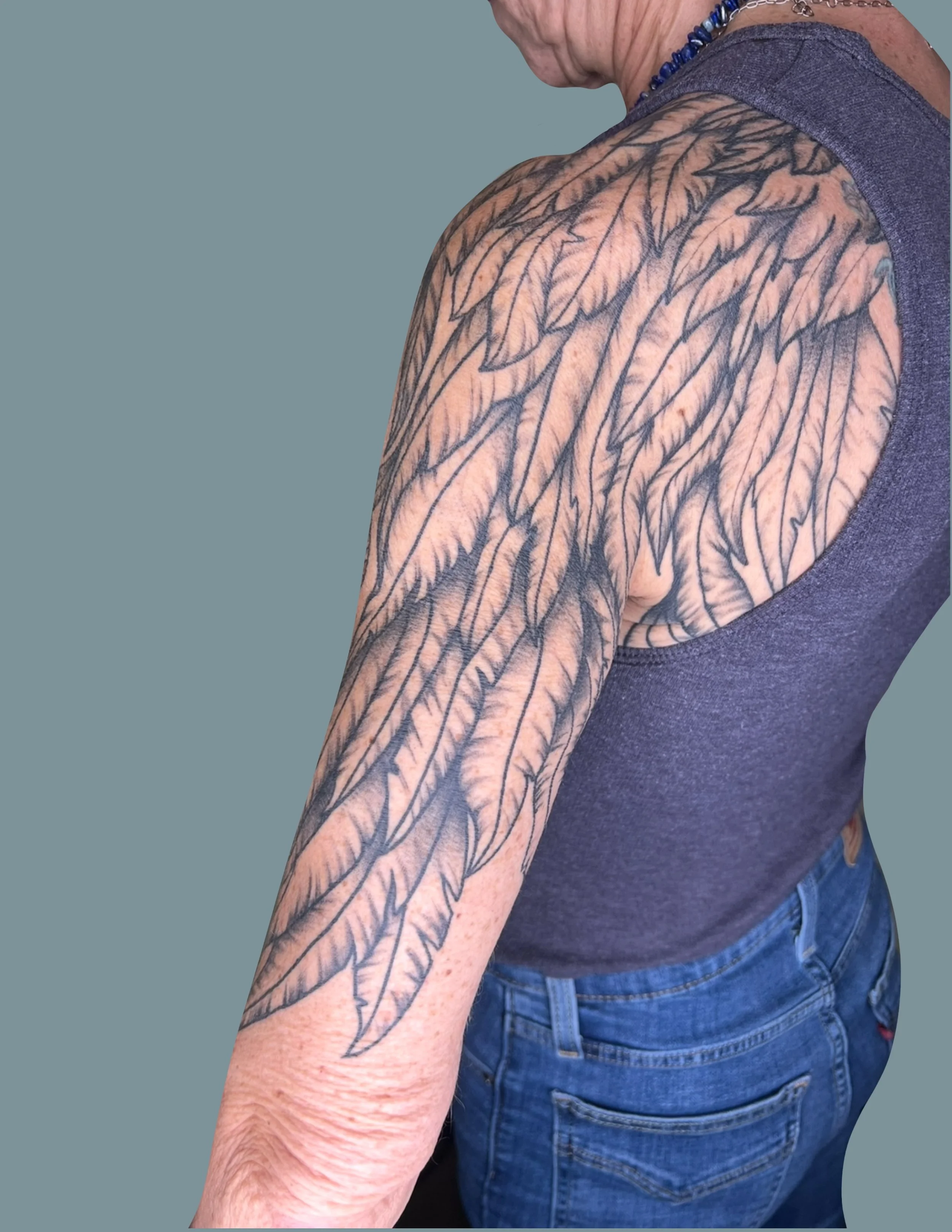 Close-up of a person's upper arm and shoulder tattoos featuring large, detailed leaves. The person is wearing a dark sleeveless shirt and blue jeans.