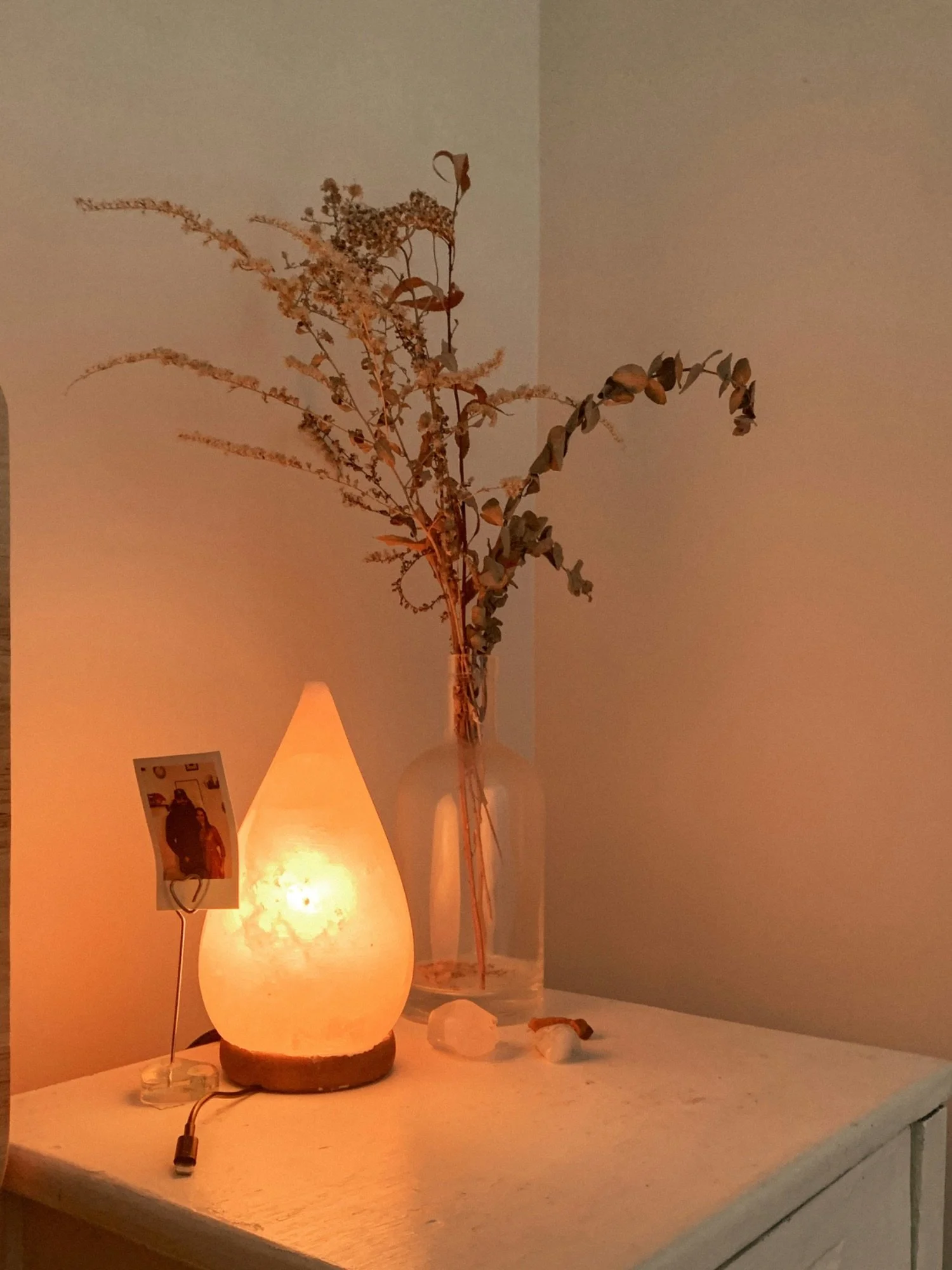 A lit Himalayan salt lamp on a white surface next to a clear vase with tall, dried, wilted plants and a small photo frame with a picture. There are also some small rocks and shells on the surface.