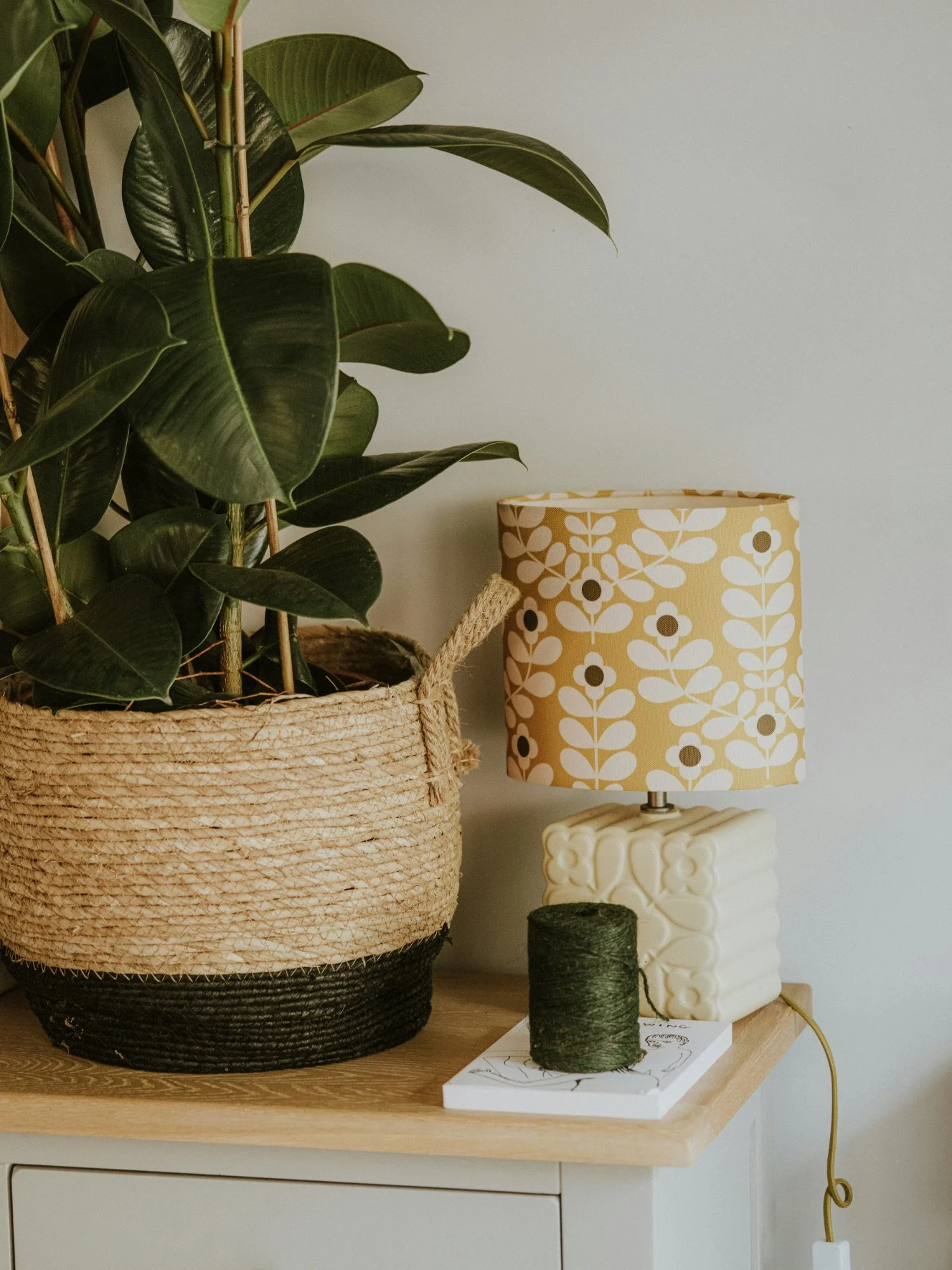 A potted plant with large green leaves in a woven basket, a yellow and white floral-patterned lampshade on a unique white base, and a small green ball of yarn on top of a piece of paper on a wooden table.