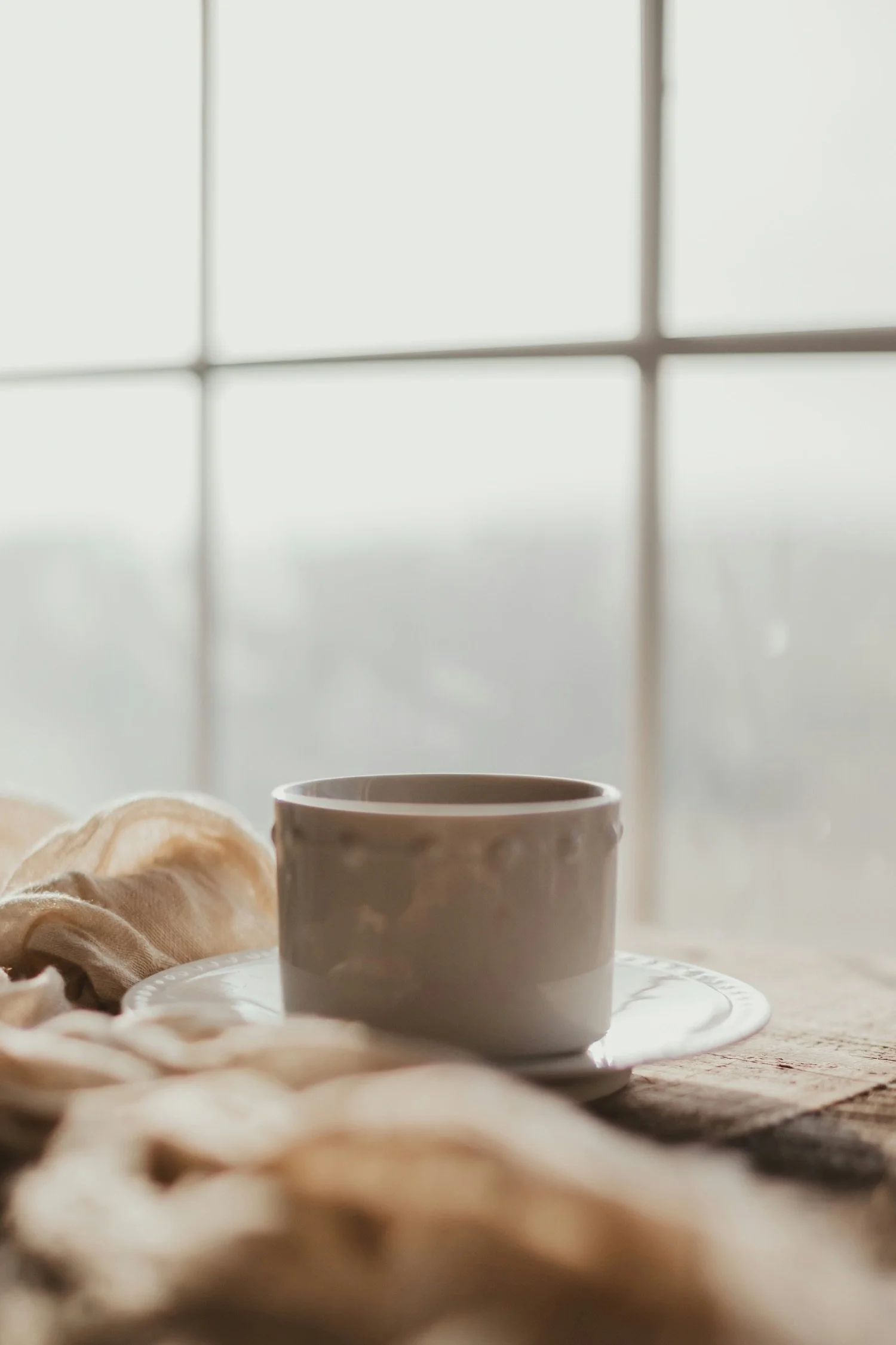 A white ceramic cup on a saucer placed on a wooden surface in front of a large window with a grid pattern, with soft bedding or fabric nearby
