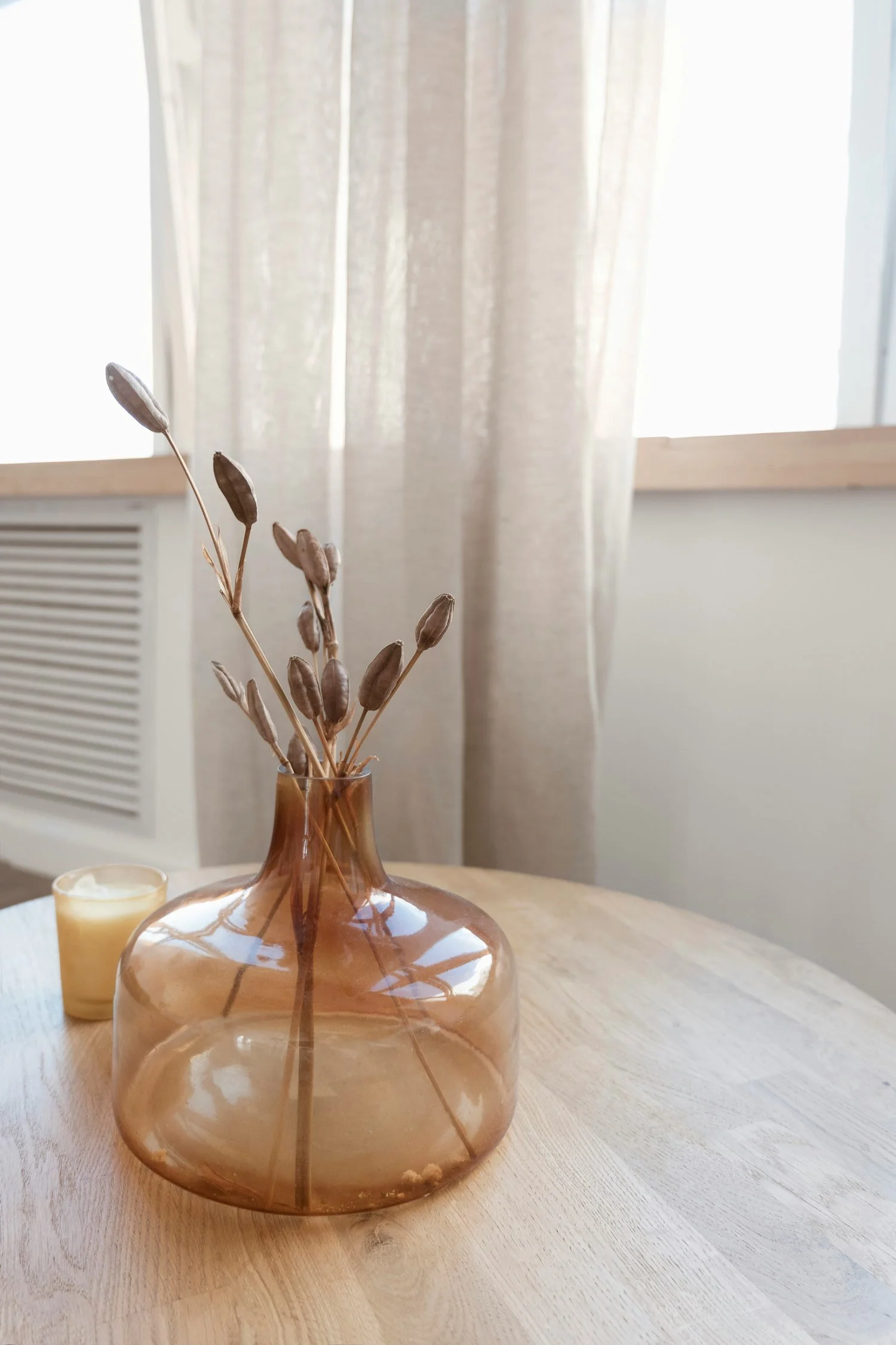 A wooden table with a tan glass vase holding dried plant stems, a small yellow candle, and a window with beige curtains in the background.