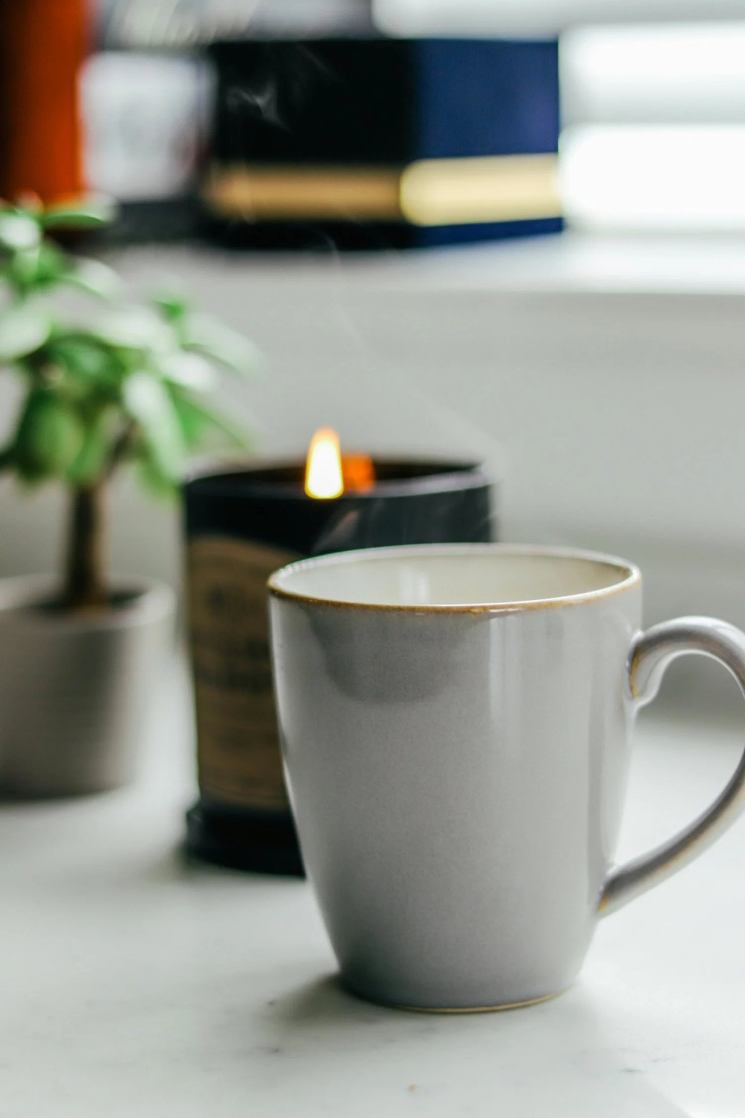 A white ceramic coffee mug on a white surface with a potted plant and a lit candle in the background.