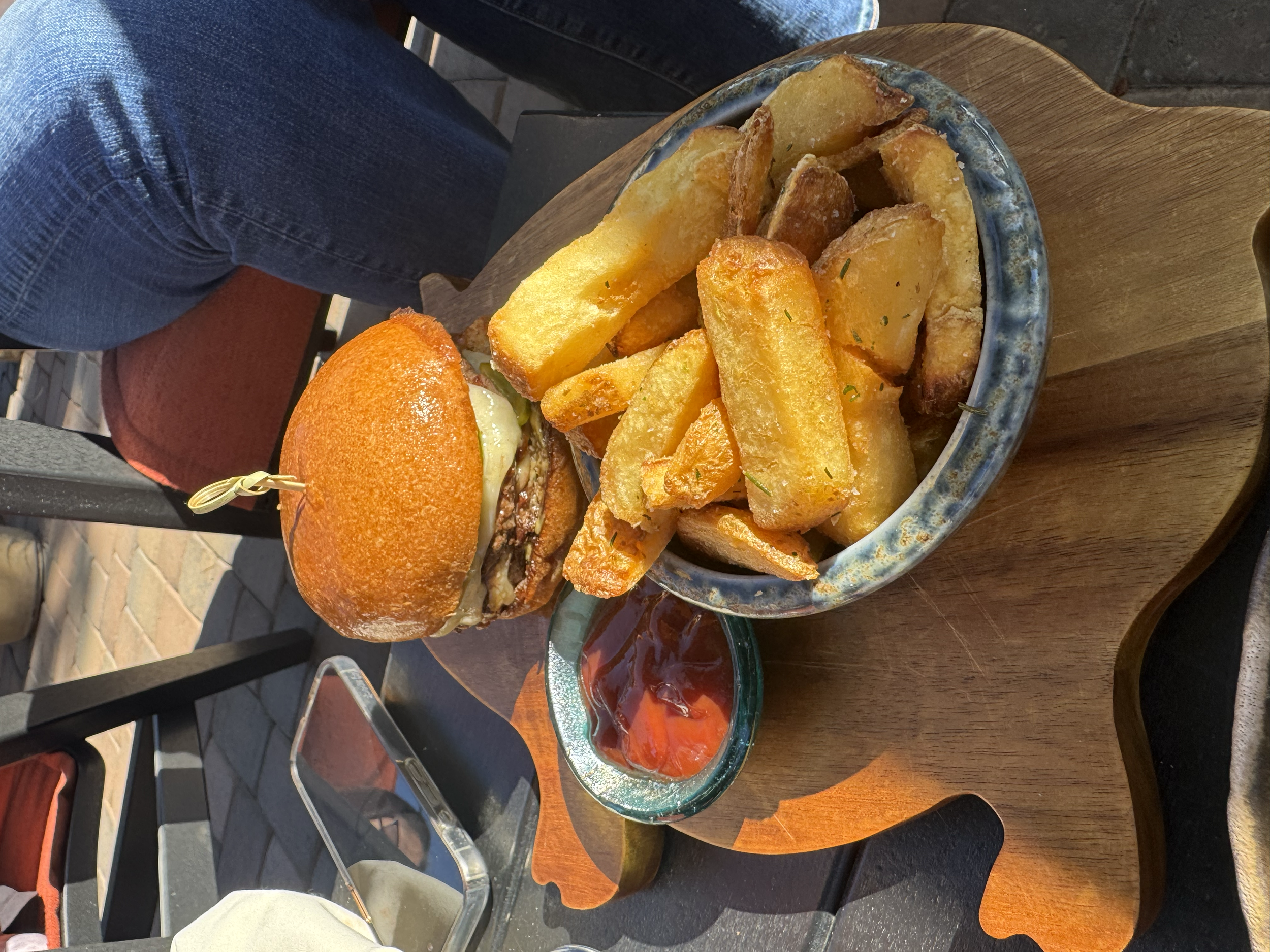 A burger with a toasted bun, melted cheese, and fillings on a wooden tray. Next to it is a bowl of thick-cut fries and a small dish of ketchup.
