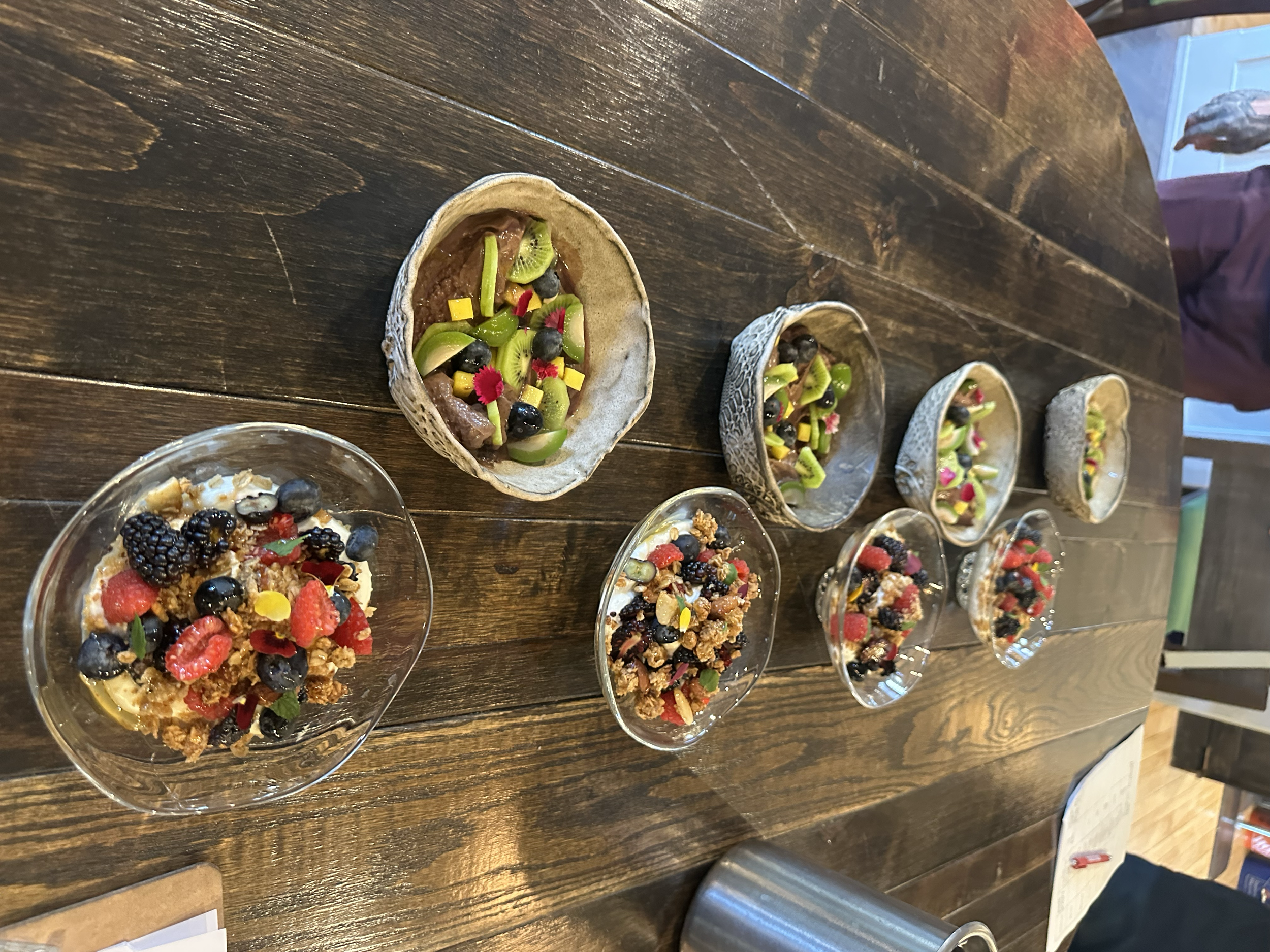 Multiple bowls of dessert with fresh berries, granola, and fruit on a wooden table.