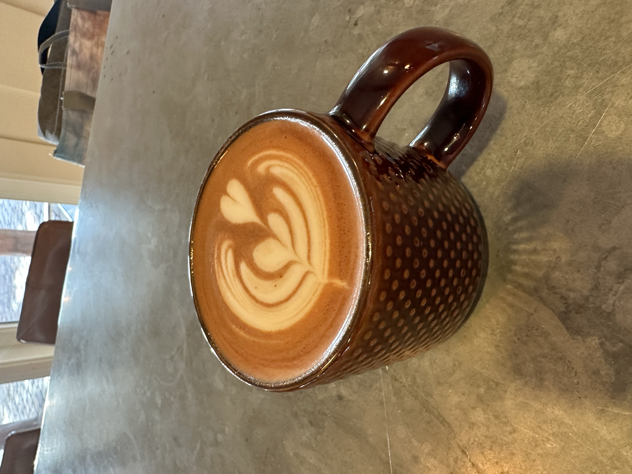 A ceramic mug with a dotted pattern, filled with a latte that has latte art in the shape of a tulip with hearts, on a beige countertop. In the background, there's a window and a chair.