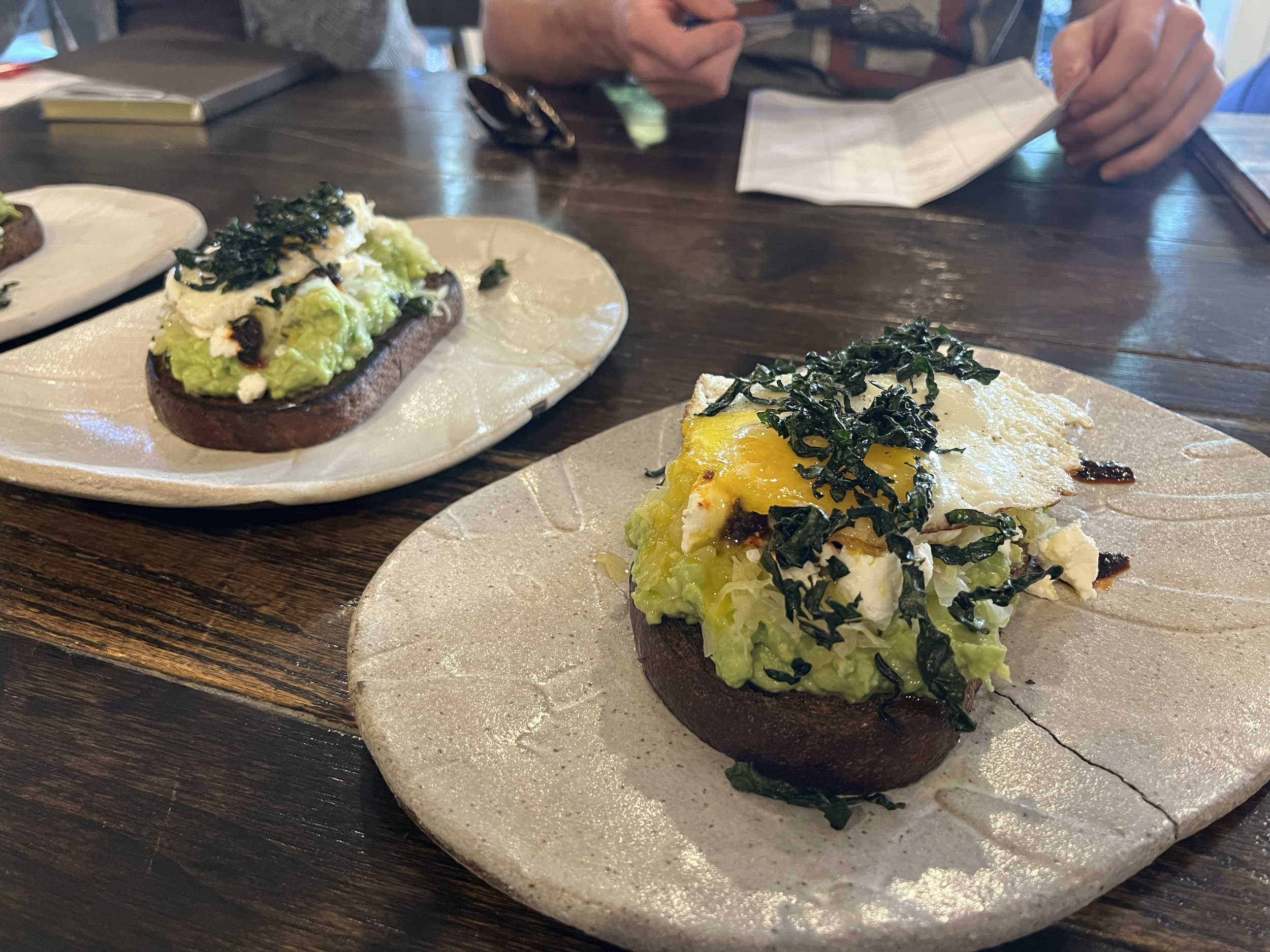 Two dishes of avocado toast topped with eggs, kale, and cheese on ceramic plates on a wooden table, with a person's hands and a map in the background.
